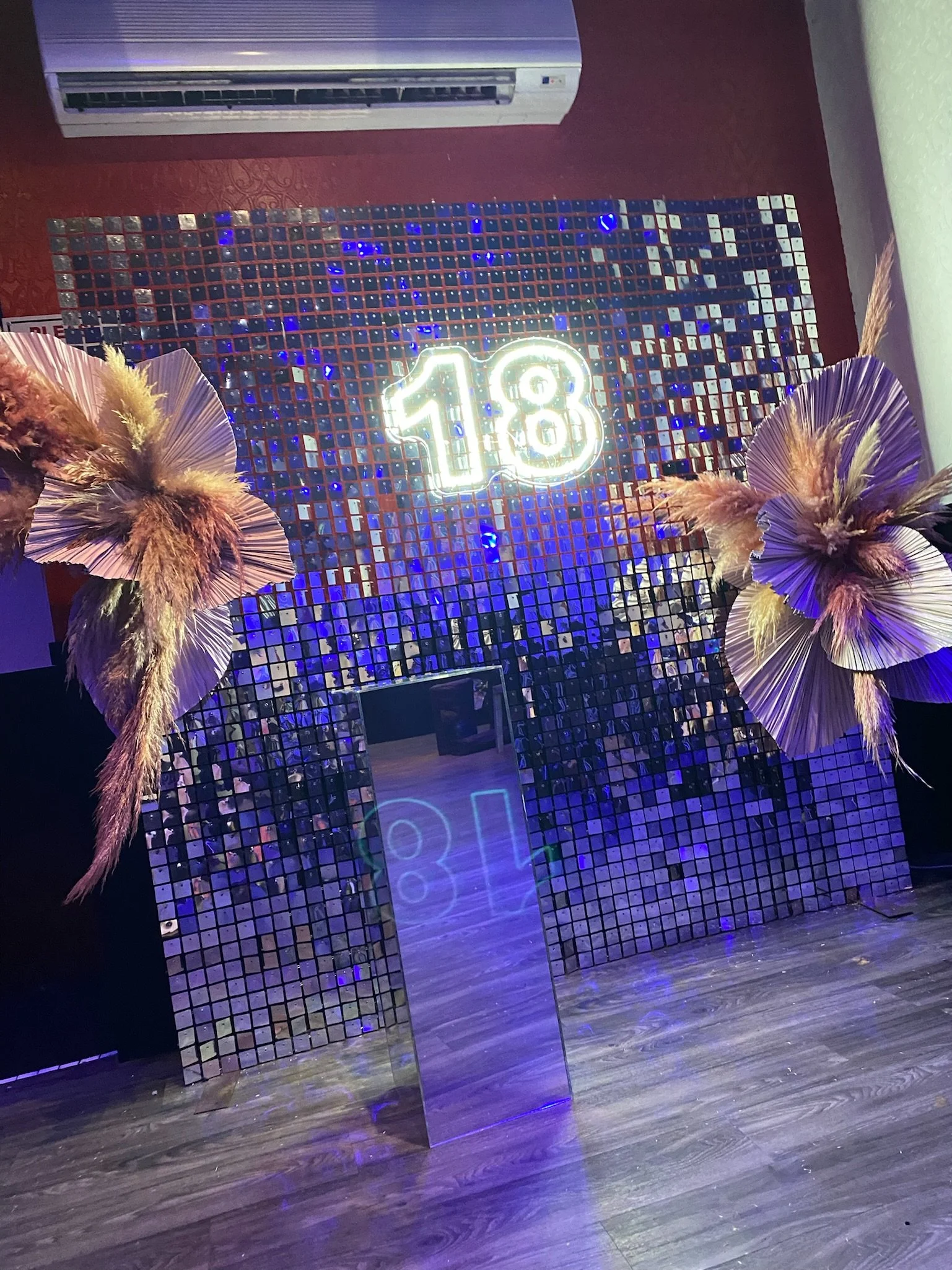 Decorative wall with small mirrored tiles, neon sign displaying '18', metallic and glass mirror panels, dried flower arrangements in pink and beige colors on either side, wooden floor, and an air conditioning unit above.