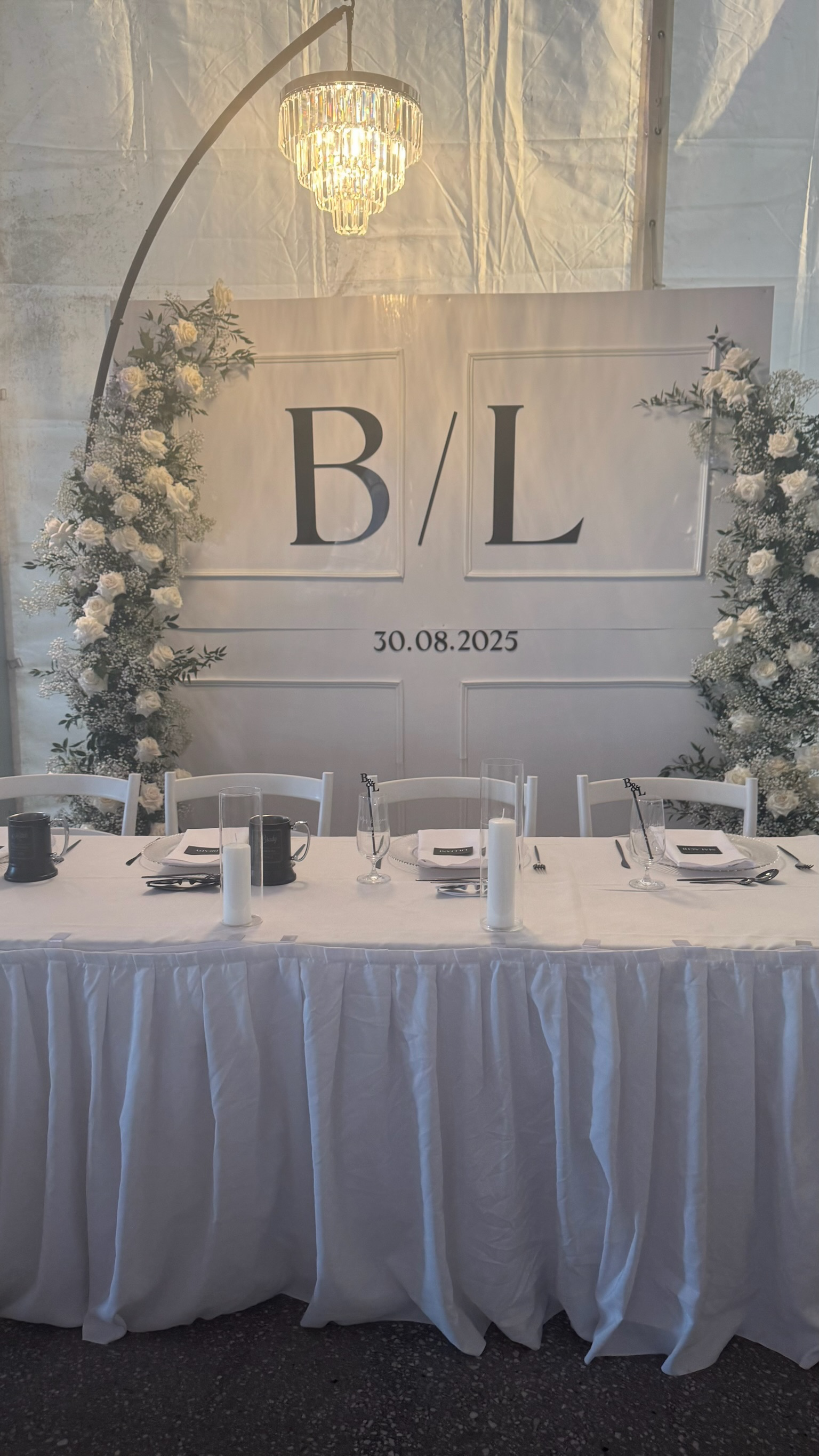 Wedding table setup with a large white backdrop featuring the initials B/L and the date 30.08.2025, decorated with white floral arrangements and candles, under a chandellier.