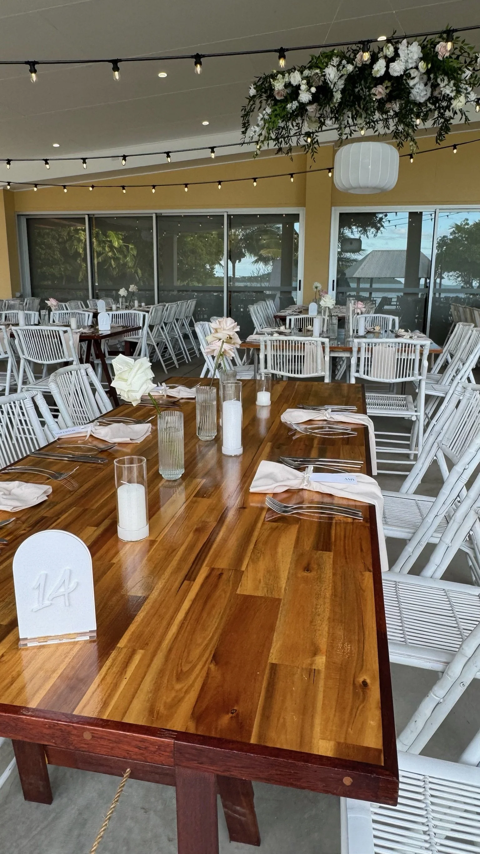 Indoor event space decorated for a gathering, with a polished wooden table set with white napkins, glass vases with flowers, and candles, surrounded by white chairs, with string lights and a large hanging floral arrangement overhead.