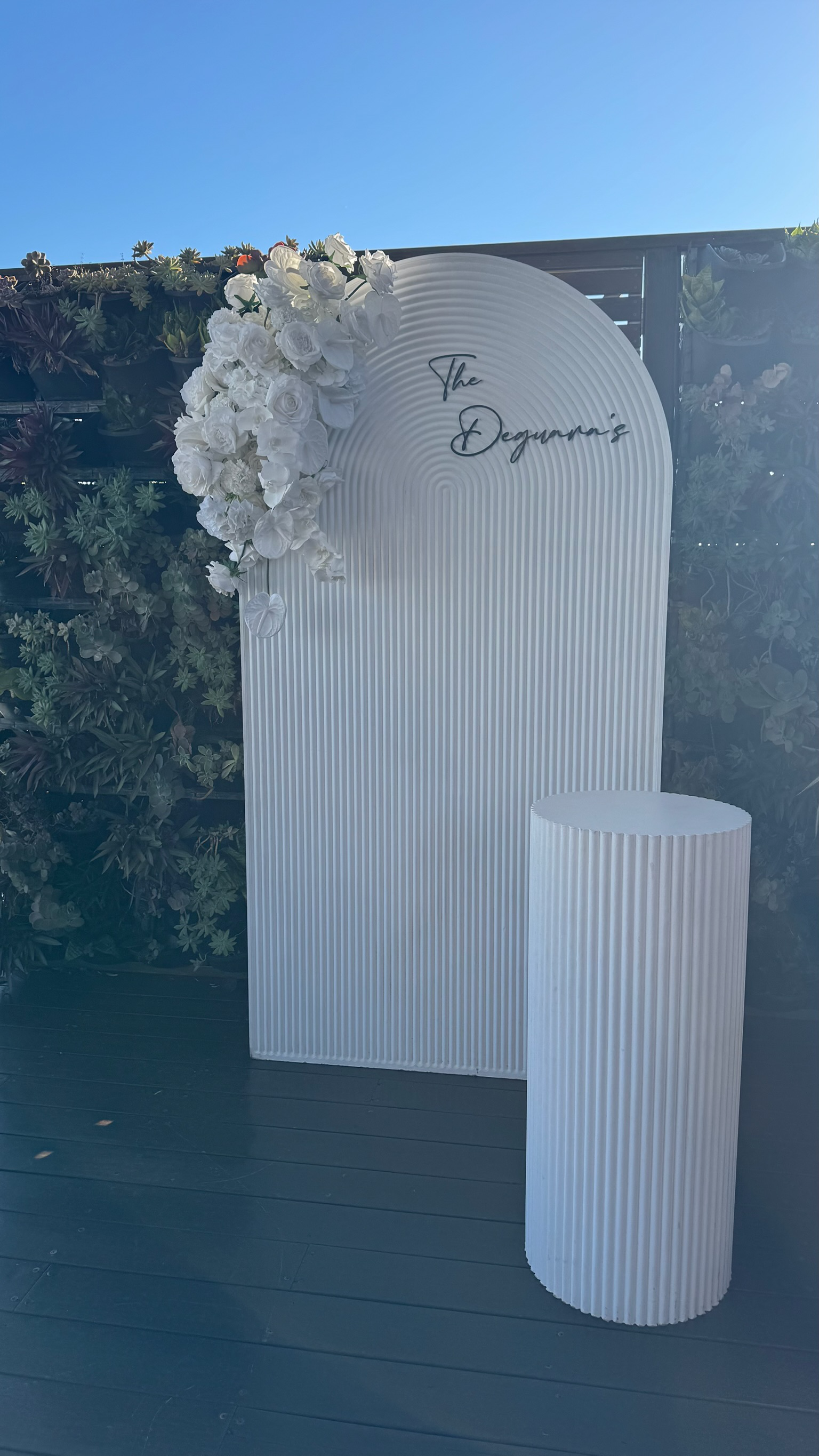 White textured backdrop with a floral decoration of white flowers on the top left corner, and the words "The Degwaras" written in script on the top right of the backdrop. A matching white cylindrical stand is placed in front.