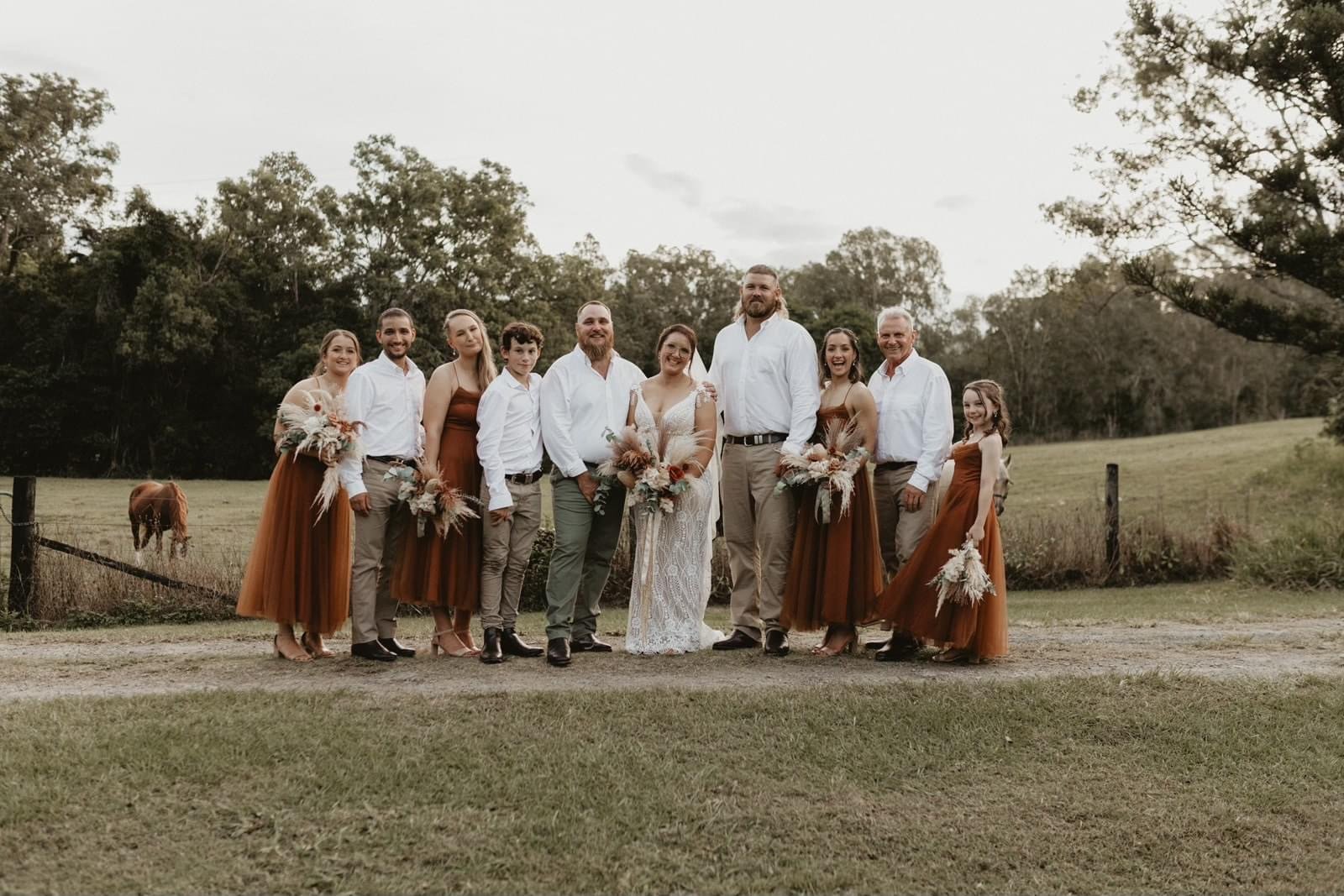 Group of people in an outdoor countryside setting, dressed in wedding and bridesmaid attire, standing on grass with trees and a horse in the background.