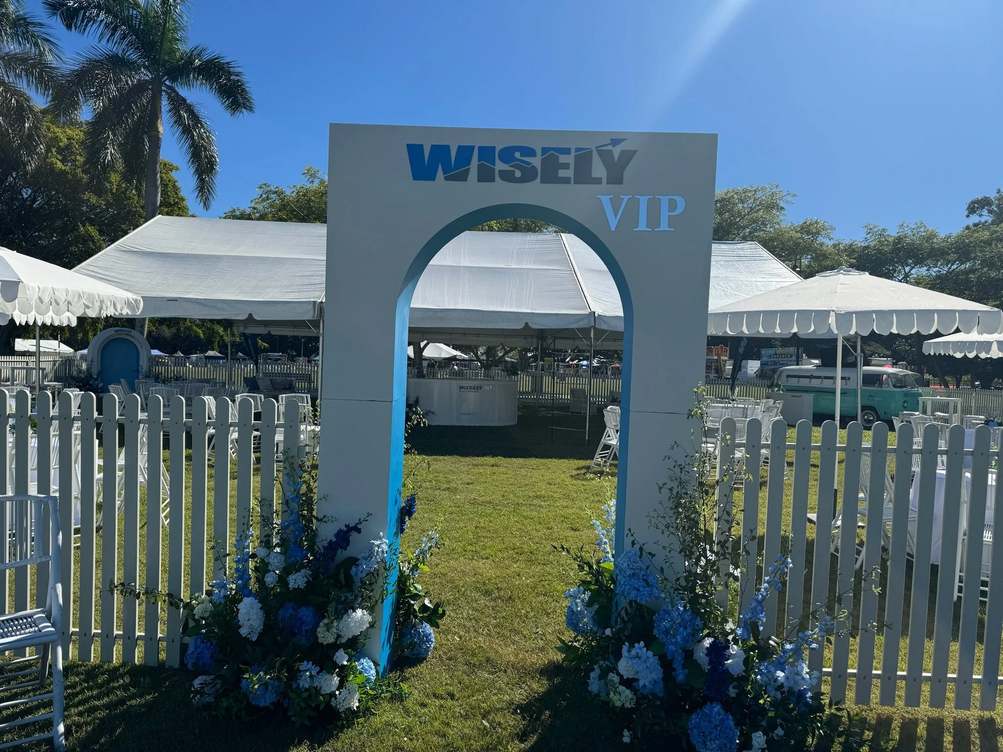 White wedding or event arch with blue and white floral arrangements, marked with 'WISLY VIP', set up outdoors with white tents, picnic tables, and white fencing under a clear blue sky.