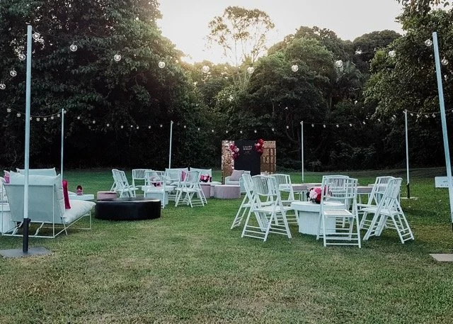 Outdoor event setup with white tables and chairs, string lights, and a decorated backdrop, on a grassy area surrounded by trees at sunset.