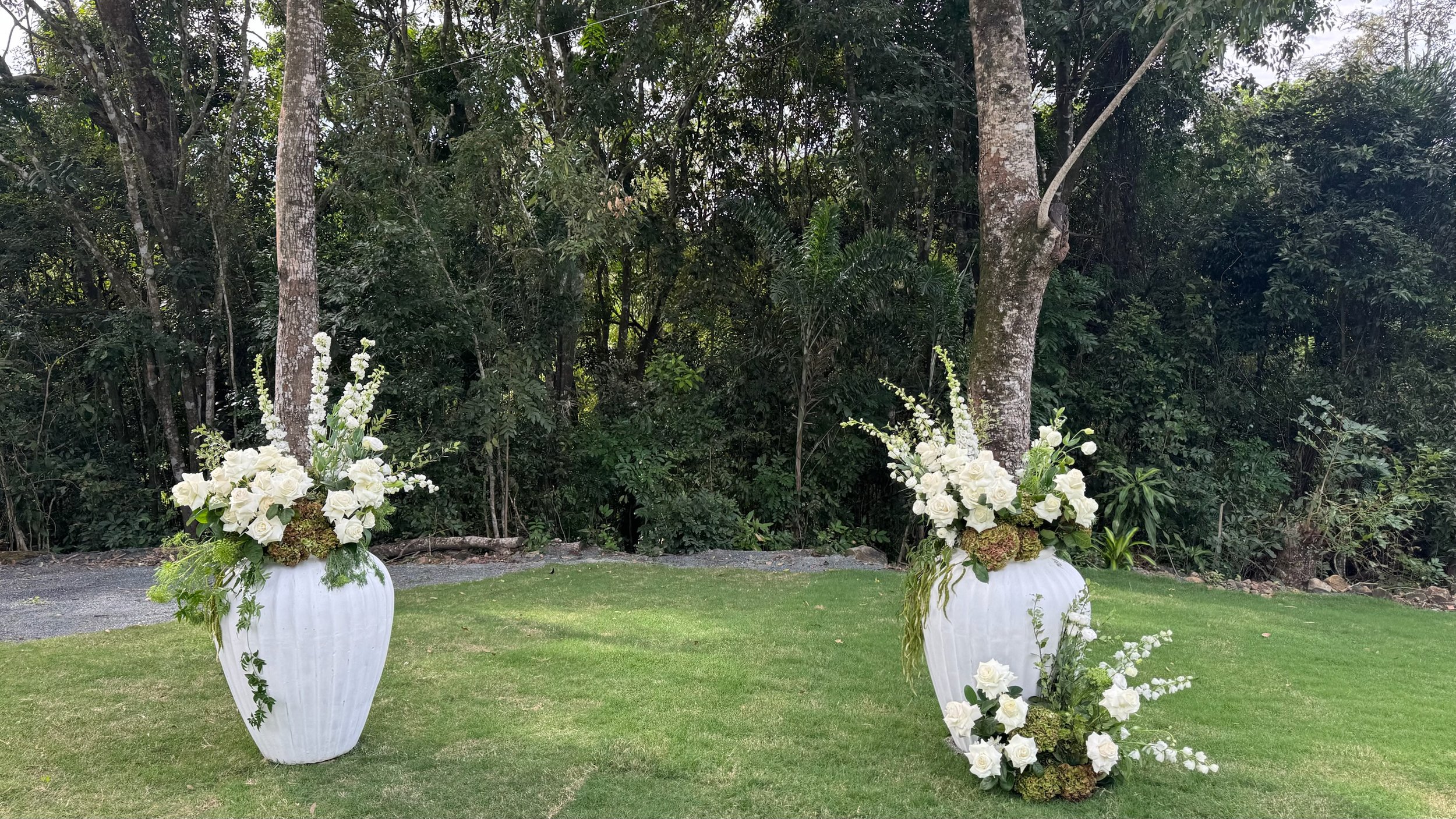 Two large white vases filled with white flowers and green foliage are placed on a grassy area in front of a wooded background, with one vase on each side and a smaller arrangement on the ground.