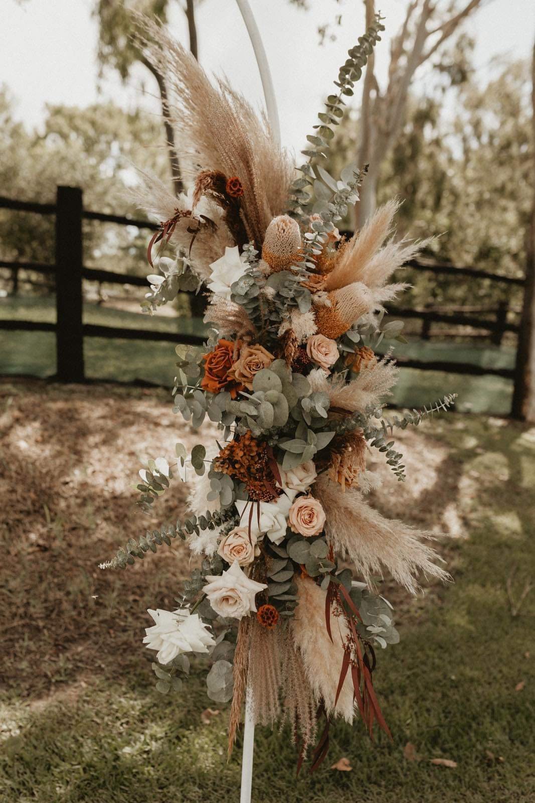 A large, vertical floral arrangement on a stand, featuring dried and fresh flowers such as roses, pampas grass, eucalyptus, and other foliage, set outdoors on grass with a wooden fence and trees in the background.