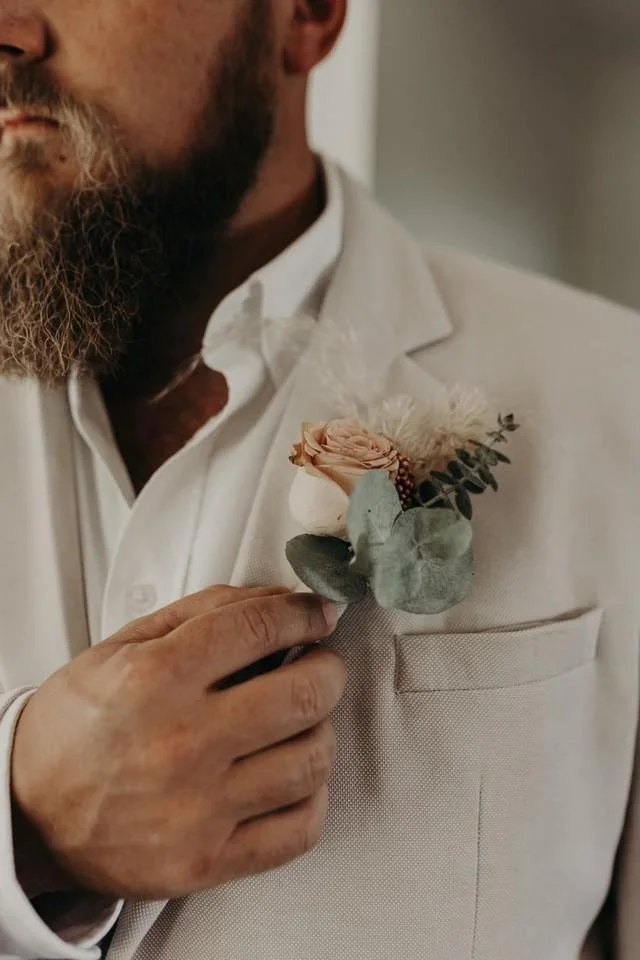 A man in a white suit holding a boutonniere with a peach-colored rose and greenery.