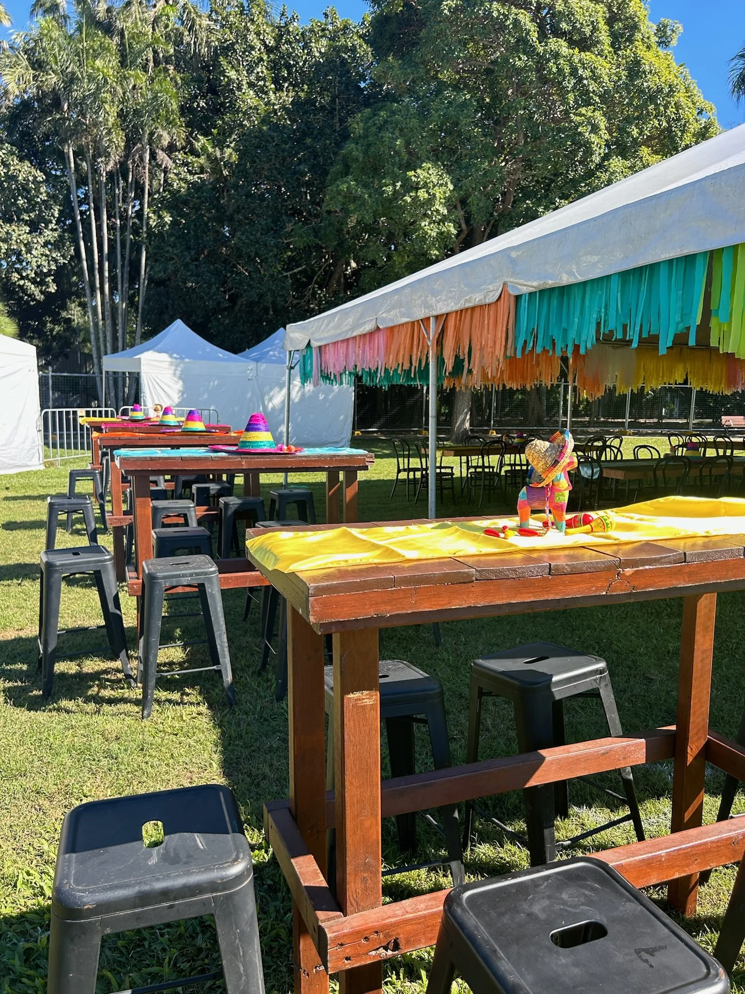 Festive outdoor setup with tables decorated for a Mexican-themed celebration, featuring colorful sombreros, a sombrero-shaped pinata, and vibrant hanging paper decorations under a tent, with lush green trees and a bright blue sky in the background.