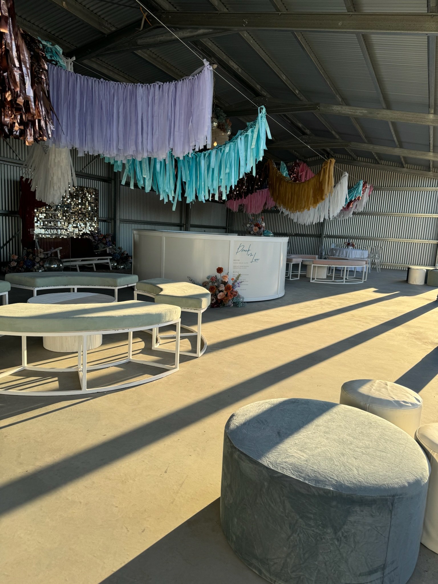 Decorative event space with colorful tissue paper tassel garlands hanging from the ceiling, white curved seating, floral arrangements, and a white reception desk in the background. The area is illuminated by natural sunlight creating shadows on the f