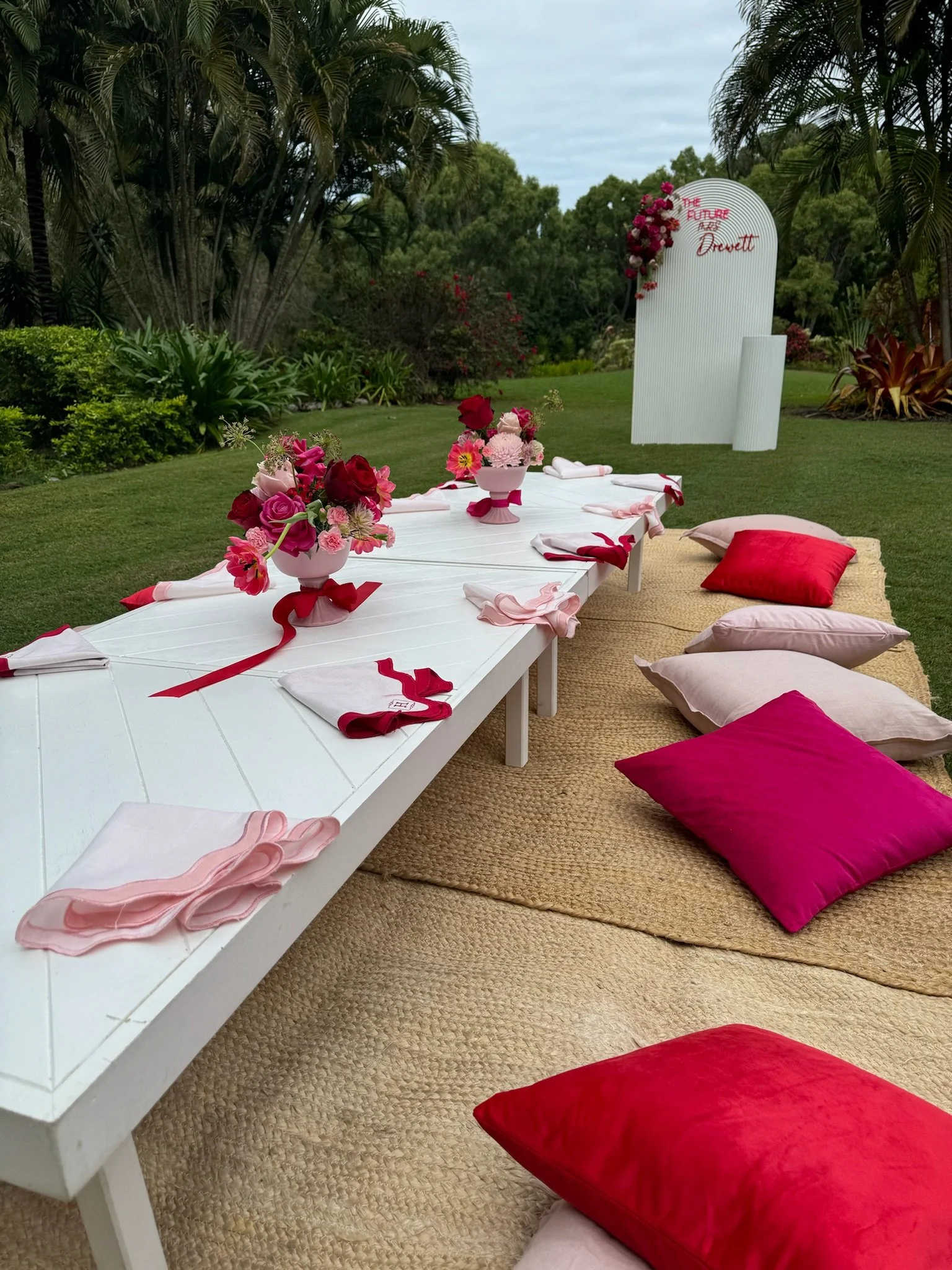 Outdoor decorated table with pink and red flower centerpieces, pink and red napkins, and colorful pillows on a beige rug, with a white backdrop reading "The Future Looks Bright" surrounded by flowers and lush greenery.