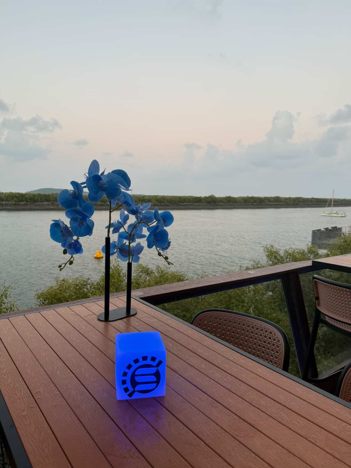 A table with a blue orchid and illuminated blue cube with black symbols, overlooking a river and a sailboat in the distance during the evening.