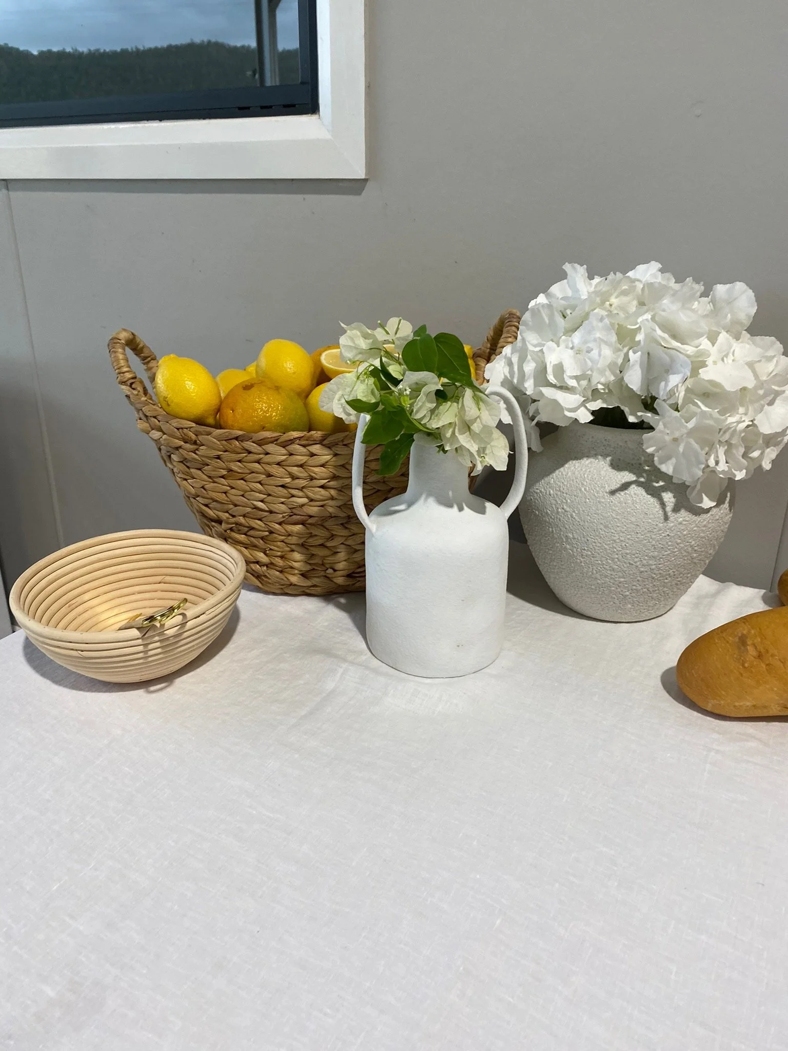 A table with a woven basket filled with lemons, a white vase with white flowers and green leaves, a large white textured pot with white flowers, and a small beige bowl containing some jewelry or keys.