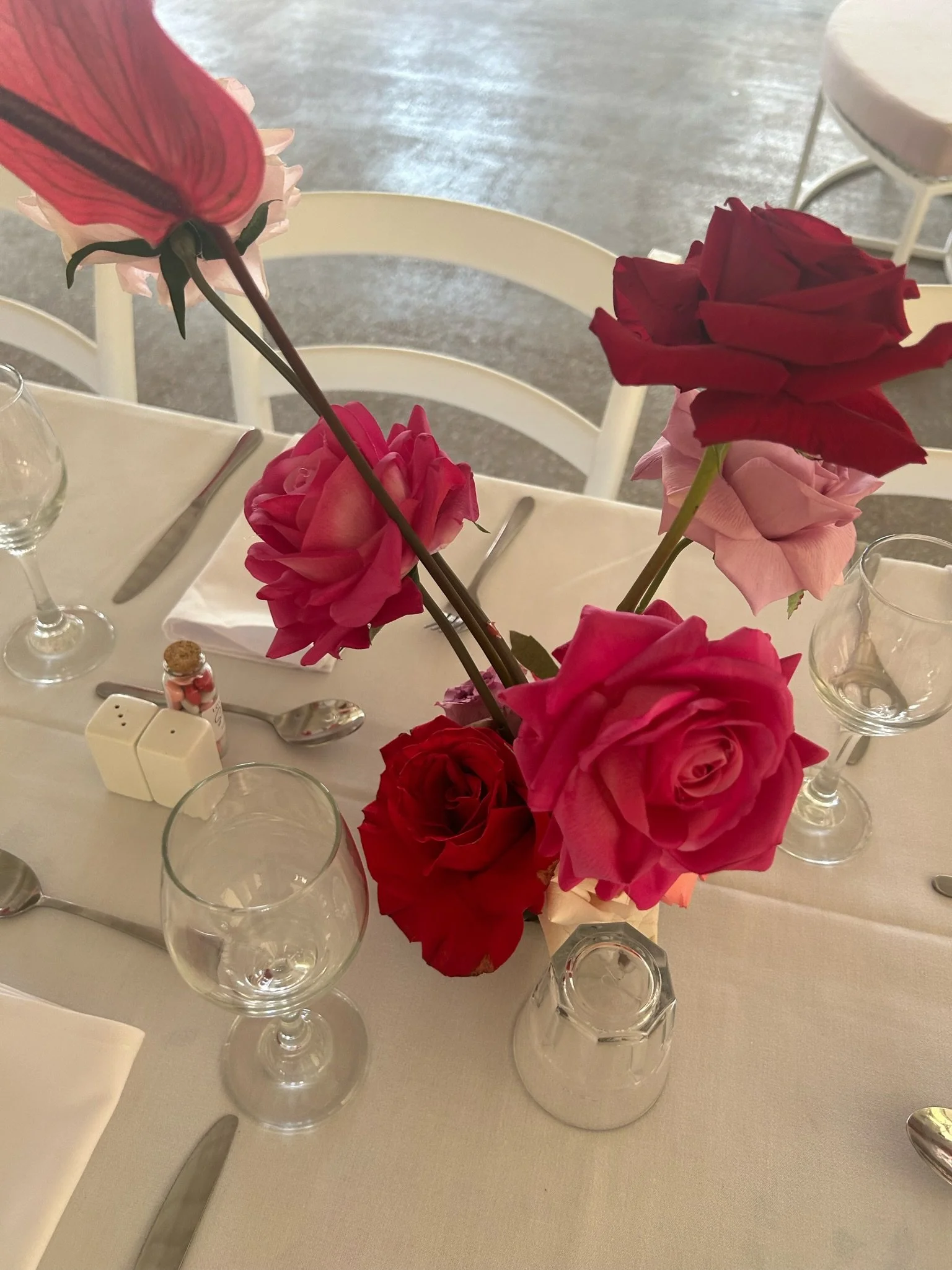 A table with a floral centerpiece of various red and pink roses, set with glassware, cutlery, and white napkins.
