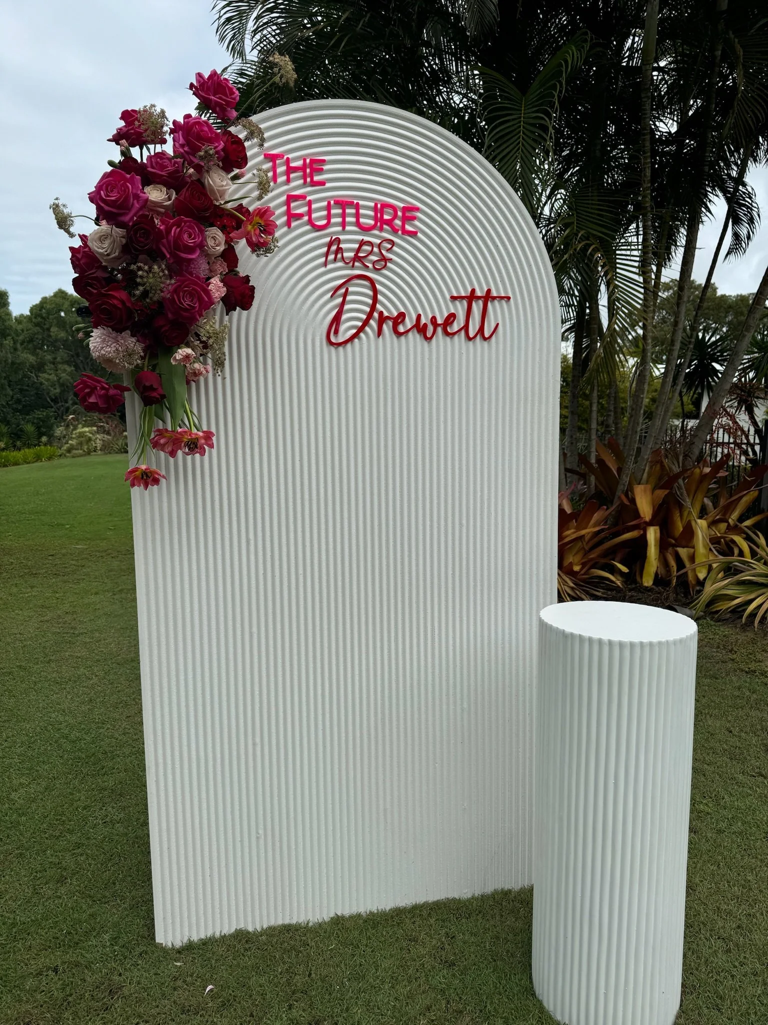 A decorative outdoor setup with a white ribbed backdrop featuring pink and white flowers on the left side. Pink text on the backdrop reads: 'THE FUTURE Mrs Drevett' in a stylish font, with 'Mrs' written in a different script.
