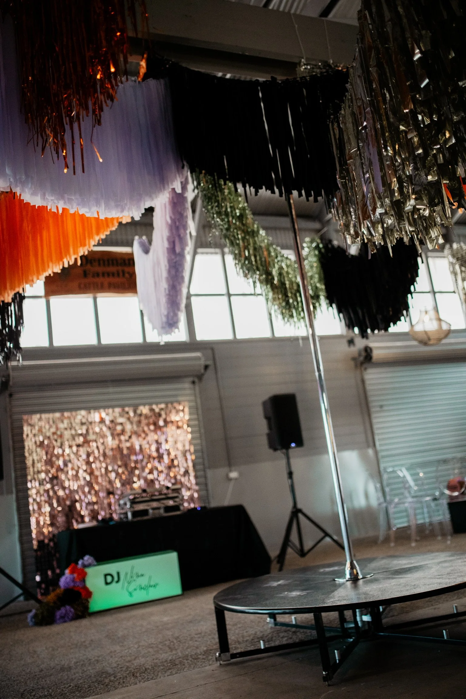 Indoor event space decorated with colorful fringed hanging decorations, a pole on a small stage, and a DJ booth in the background, suggesting a celebration or party.