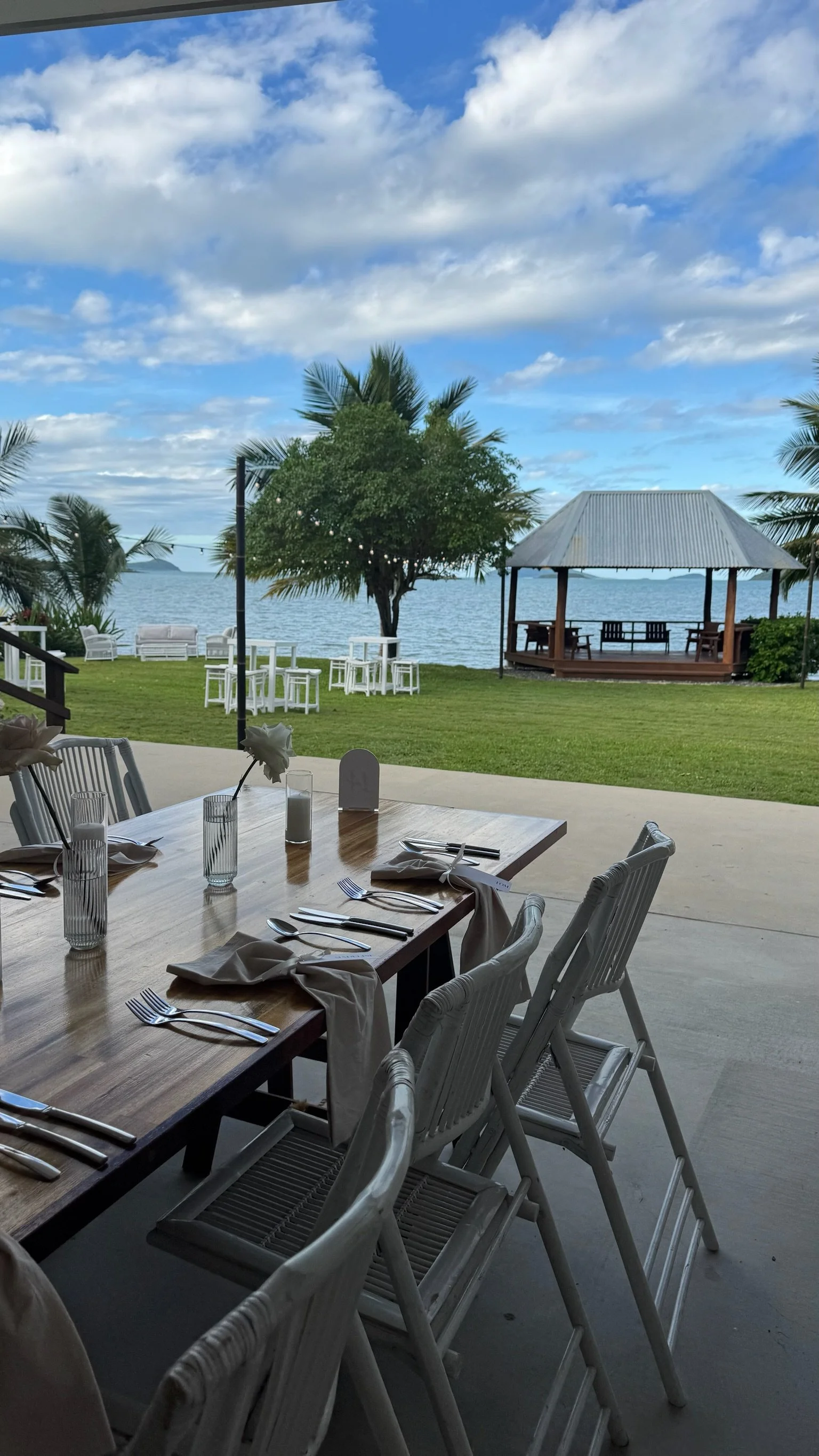 Dining area with a wooden table set with napkins, forks, knives, glasses, and a small flower arrangement, overlooking a lawn with palm trees and a gazebo by the water under a partly cloudy sky.