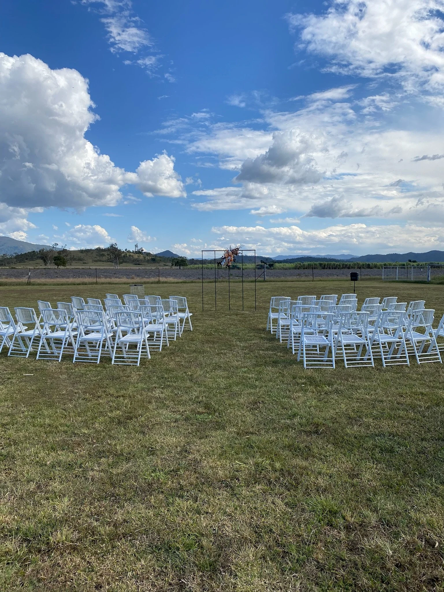 Outdoor wedding ceremony setup on a grassy field with white chairs arranged in rows facing an arch decorated with flowers, under a partly cloudy sky with mountains in the distance.