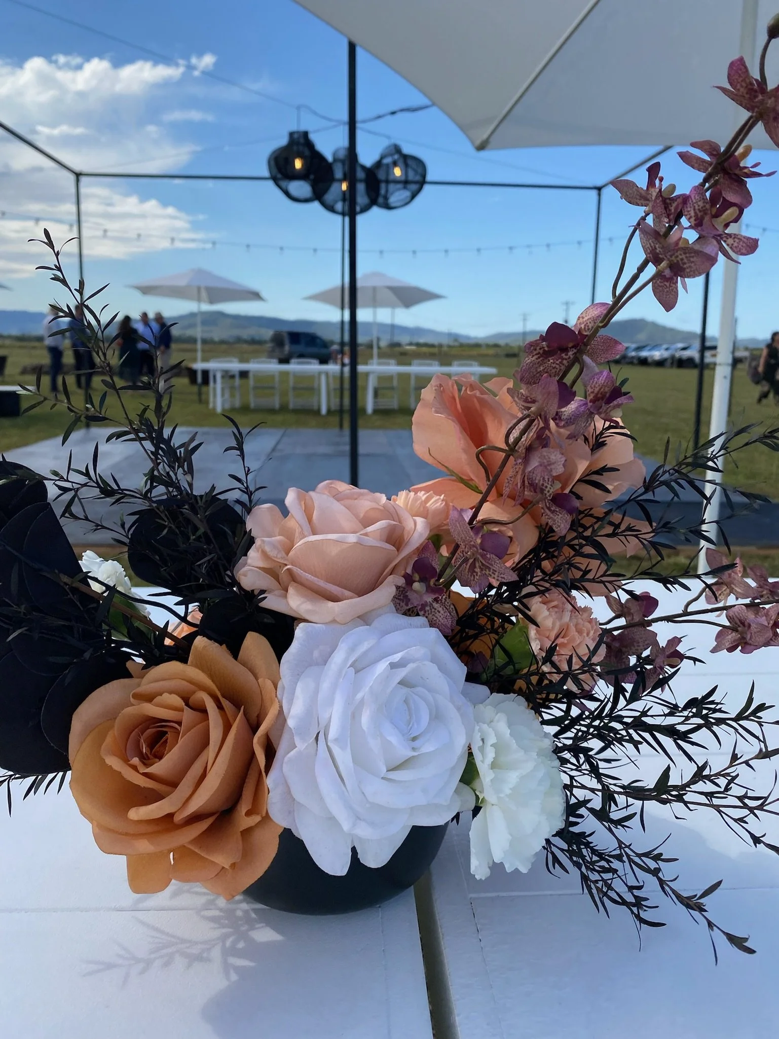 Flower arrangement with roses and greenery in a black vase, set on a table outdoors under a white canopy with string lights and a view of a field with tables, umbrellas, and distant mountains.