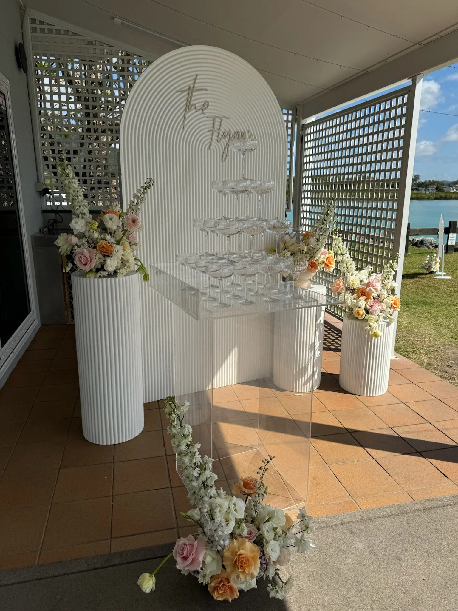 Champagne tower setup with floral arrangements on a white, ribbed table and backdrop, outdoor setting near water.