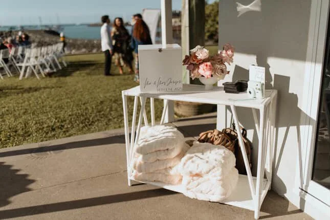 A white table with flowers and decor near a glass wall, with blankets underneath, and people in the background outdoors near water.