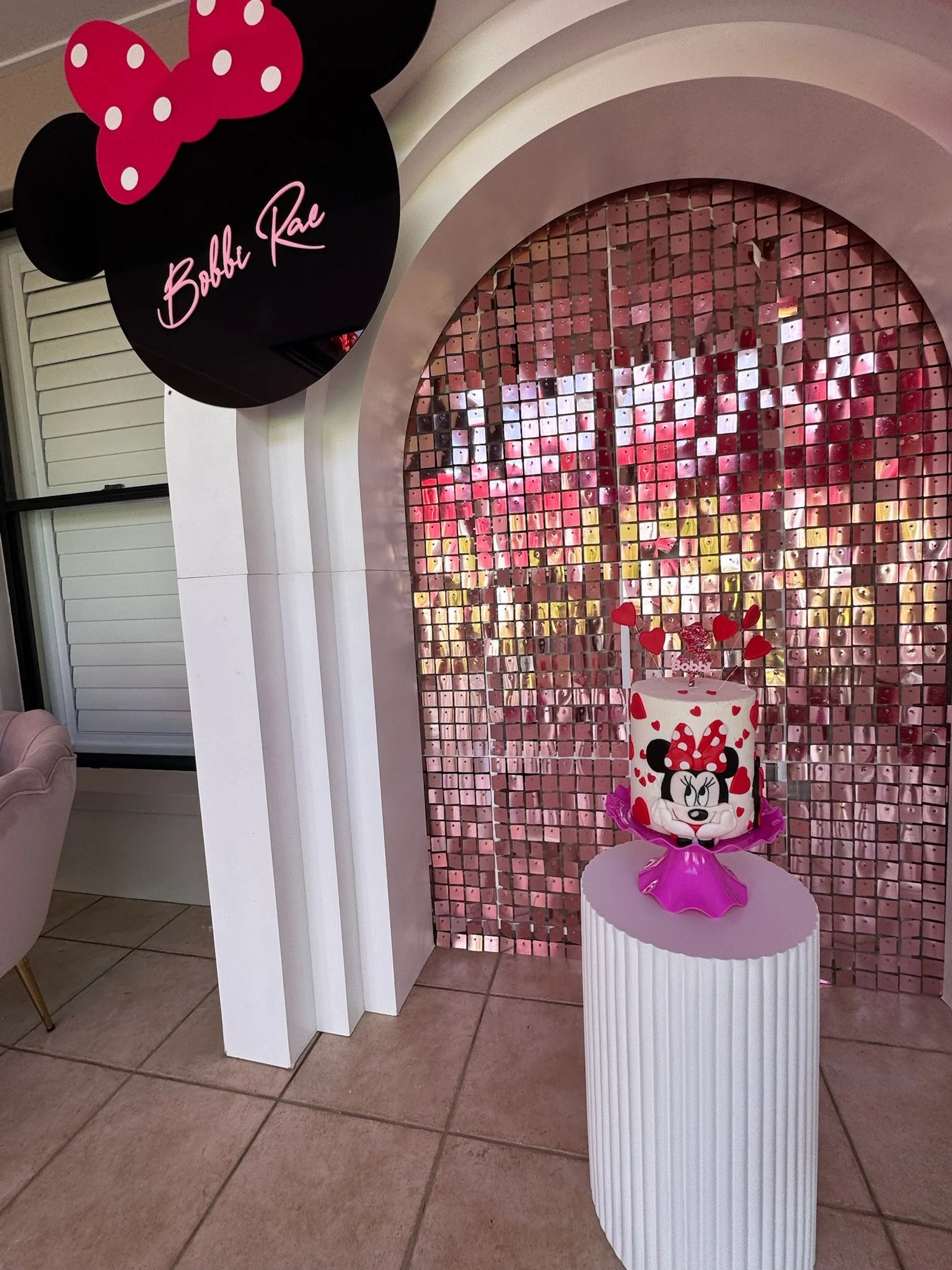 Minnie Mouse themed birthday cake with red hearts and Minnie Mouse face, pink and purple decorations, pink mirror mosaic backdrop, and "Bobbi Rae" sign with Minnie Mouse ears and bow