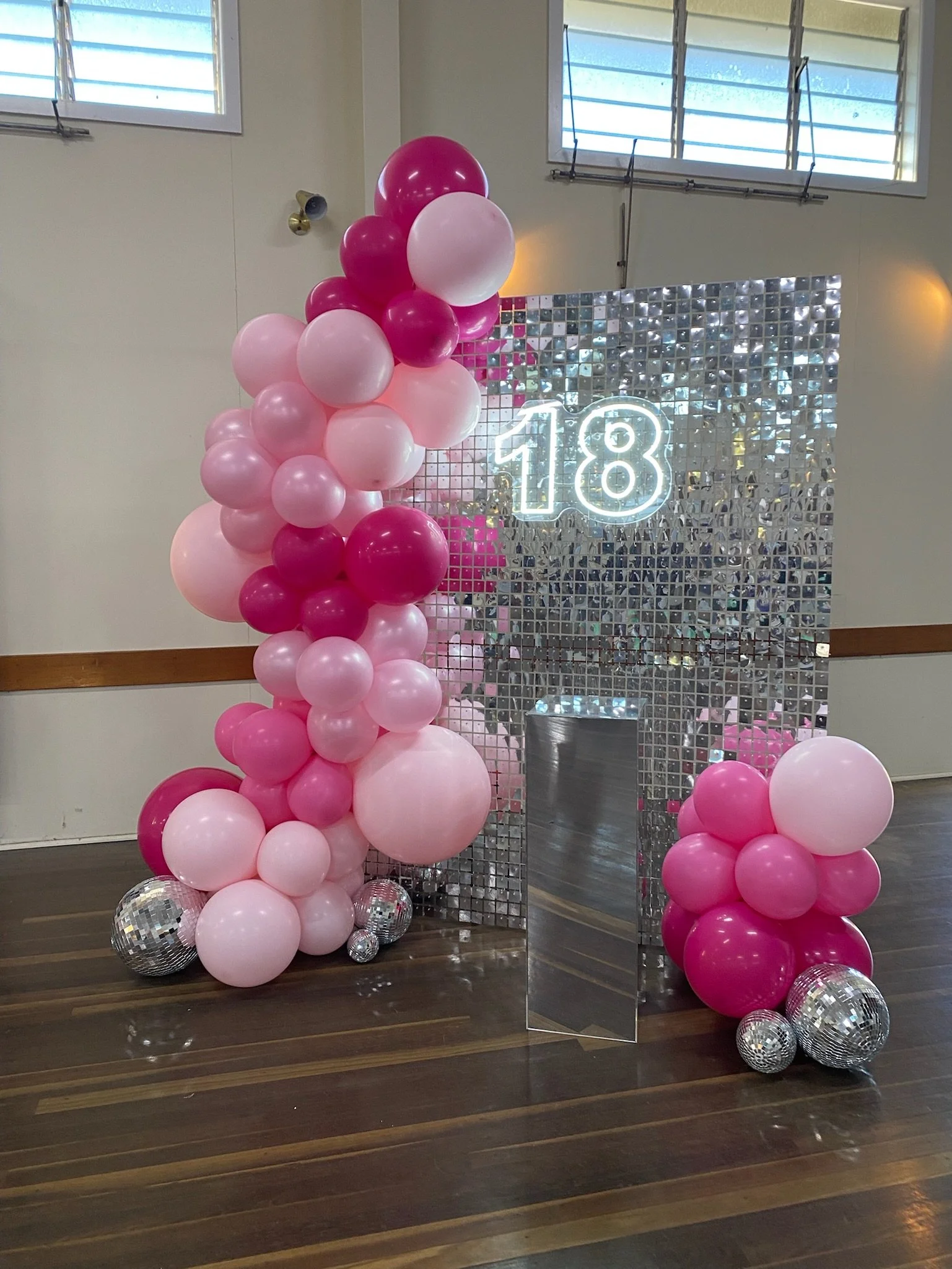 Decorative setup with pink and white balloons, a mirror panel, and a neon '18' sign for an 18th birthday celebration.