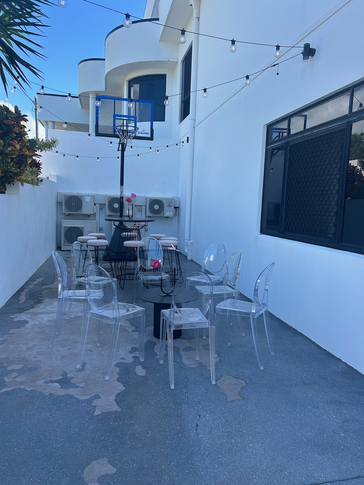 An outdoor patio area with clear plastic chairs around small black tables, a basketball hoop mounted on the wall, string lights hanging overhead, and a white building in the background.