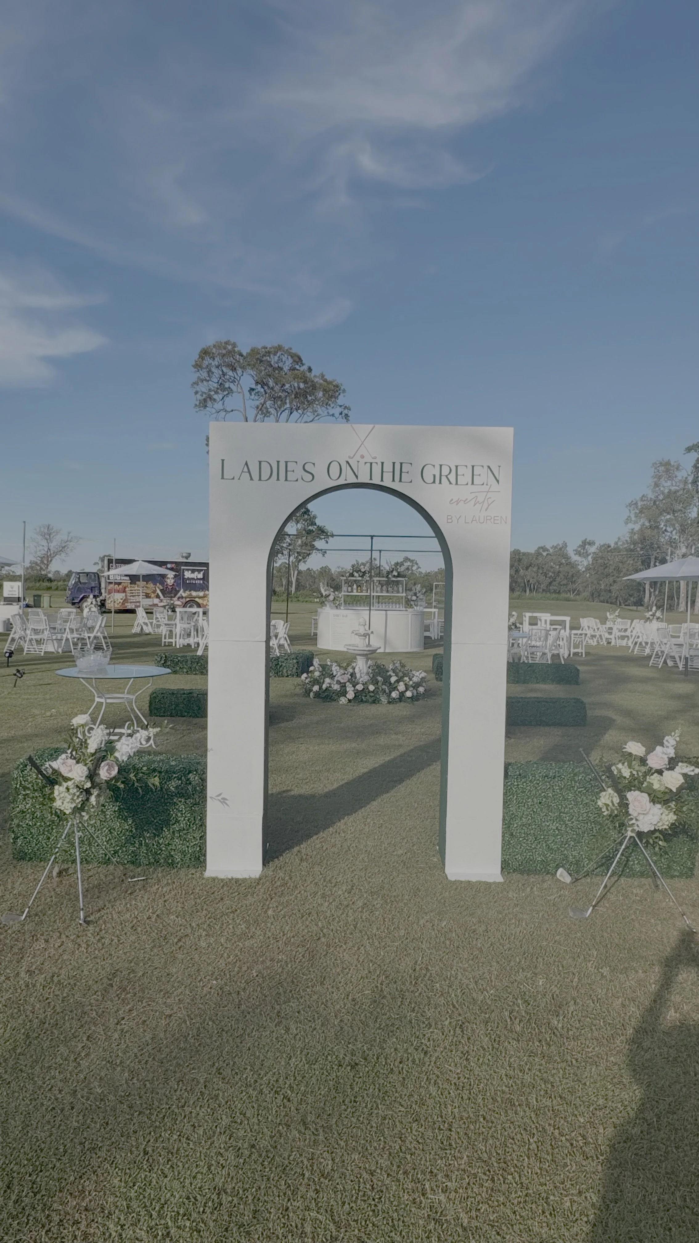 Outdoor wedding or event setup with a white arch labeled "Ladies on the Green," surrounded by flowers and decorated tables on a grassy field under a blue sky.