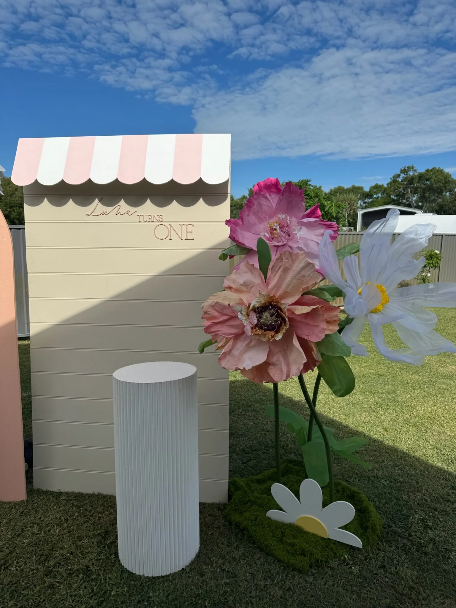 Decorative outdoor display with a large flower sculpture, a small rectangular white freestanding display with vertical lines, and a backdrop resembling a small storefront with pink and white striped awning and the words "Luha turns ONE"