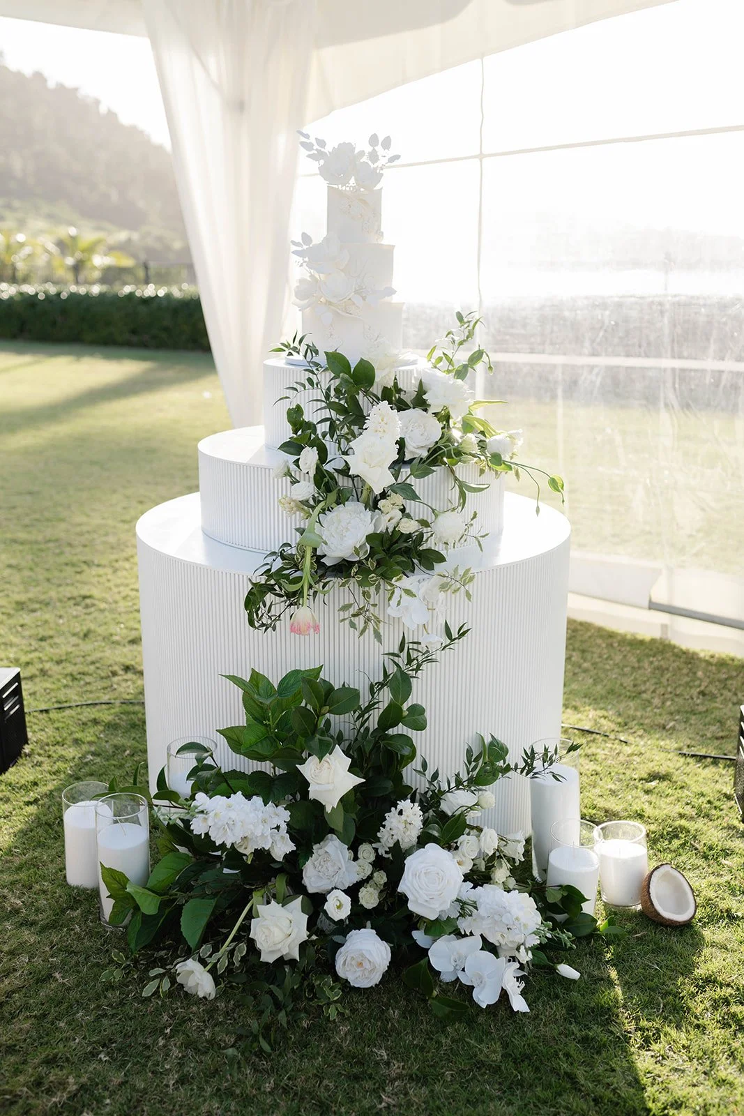 White wedding cake with cascading white flowers and green foliage, surrounded by white candles on a grassy area in a bright outdoor setting.