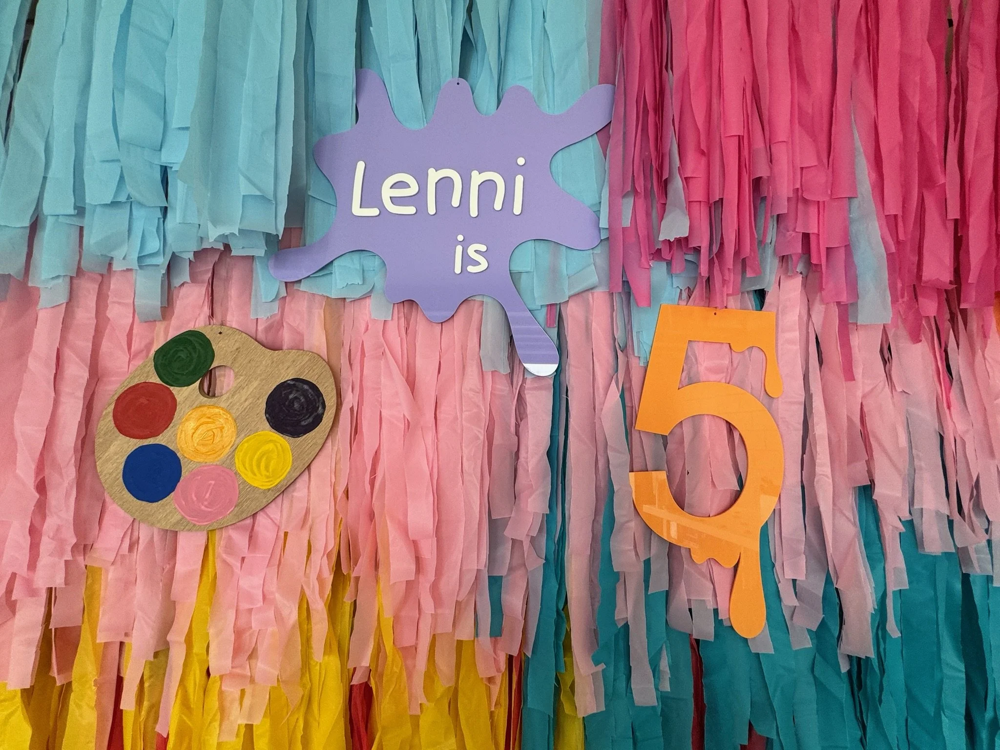Decorative backdrop with pastel-colored tissue paper strips, a purple sign that says 'Lenni is', a paper artist palette with paint colors, and large orange cutout numbers '5' and '0' indicating a 50th celebration.