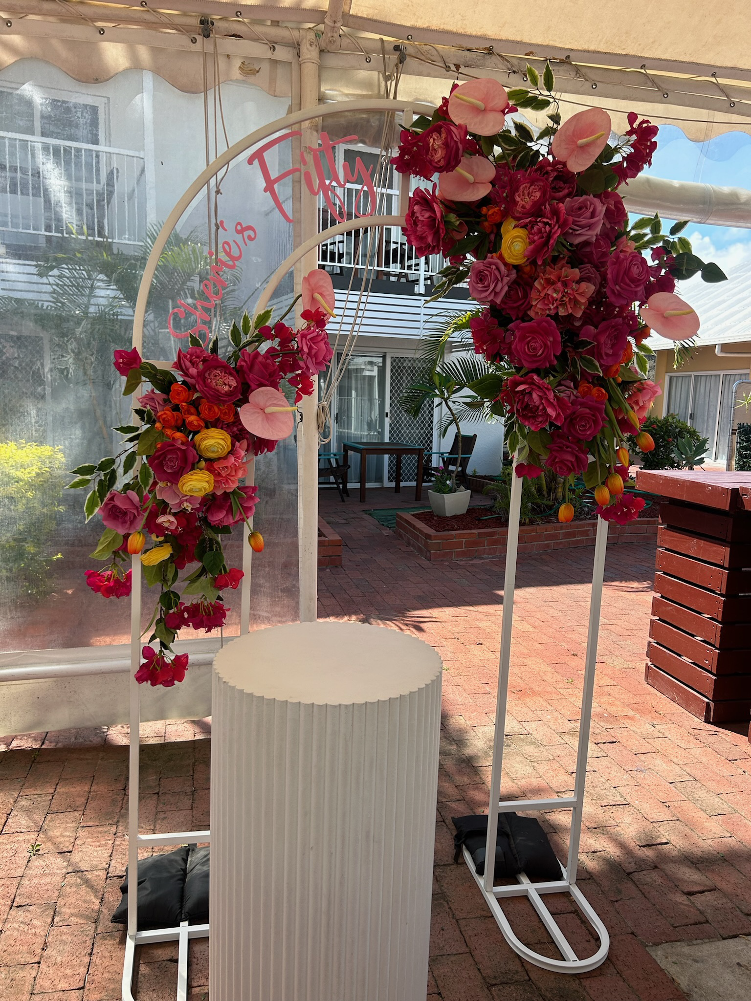 A floral arch with pink, red, orange, and yellow flowers, and a sign that reads 'Shenice's Fitties'.