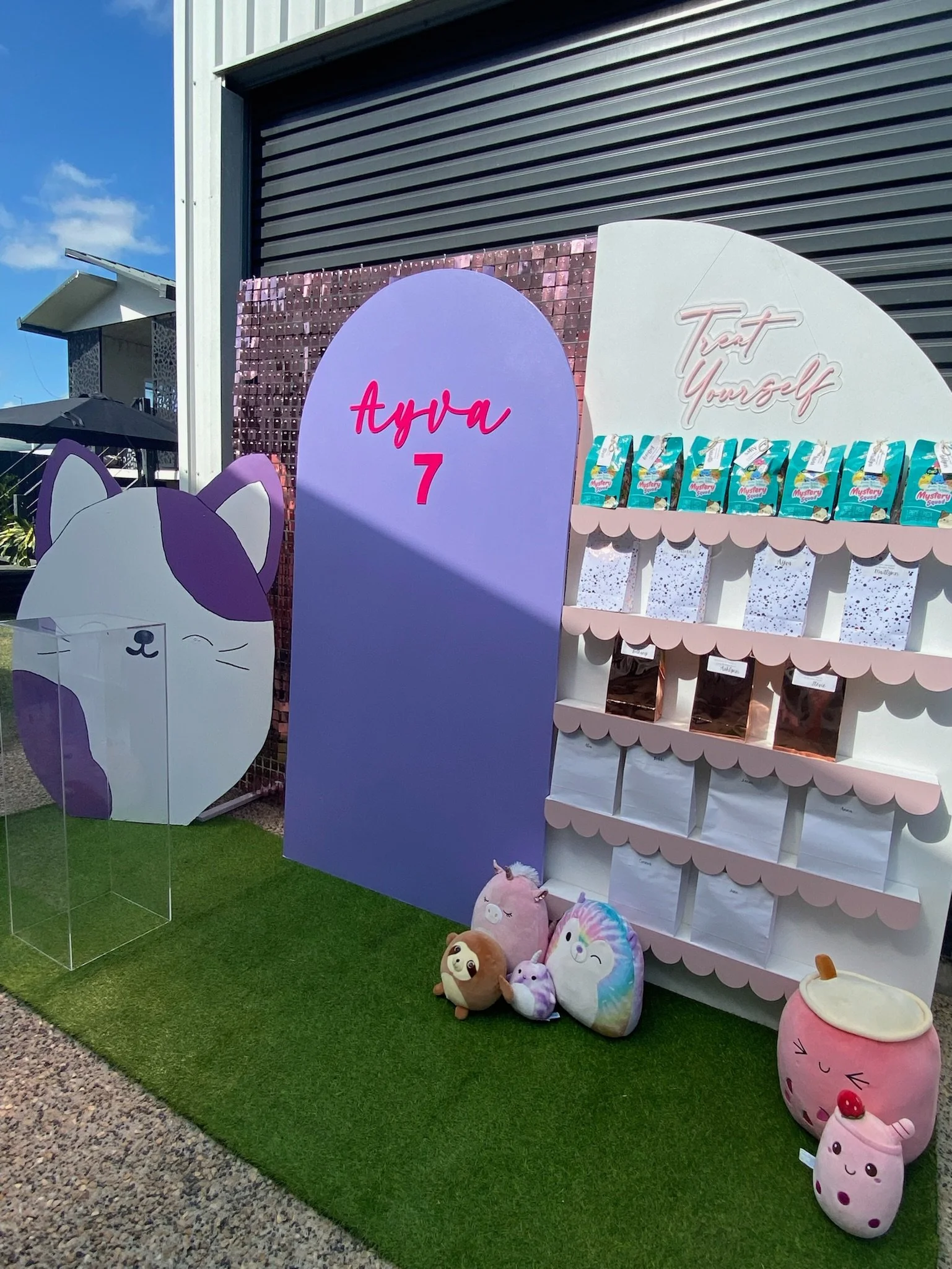 Colorful display setup with plush toys, a purple backdrop that says 'Aya 7', a white and pink shelving unit, and a large cartoon cat face with a purple ear on the left. Green turf on the ground and an umbrella in the background.