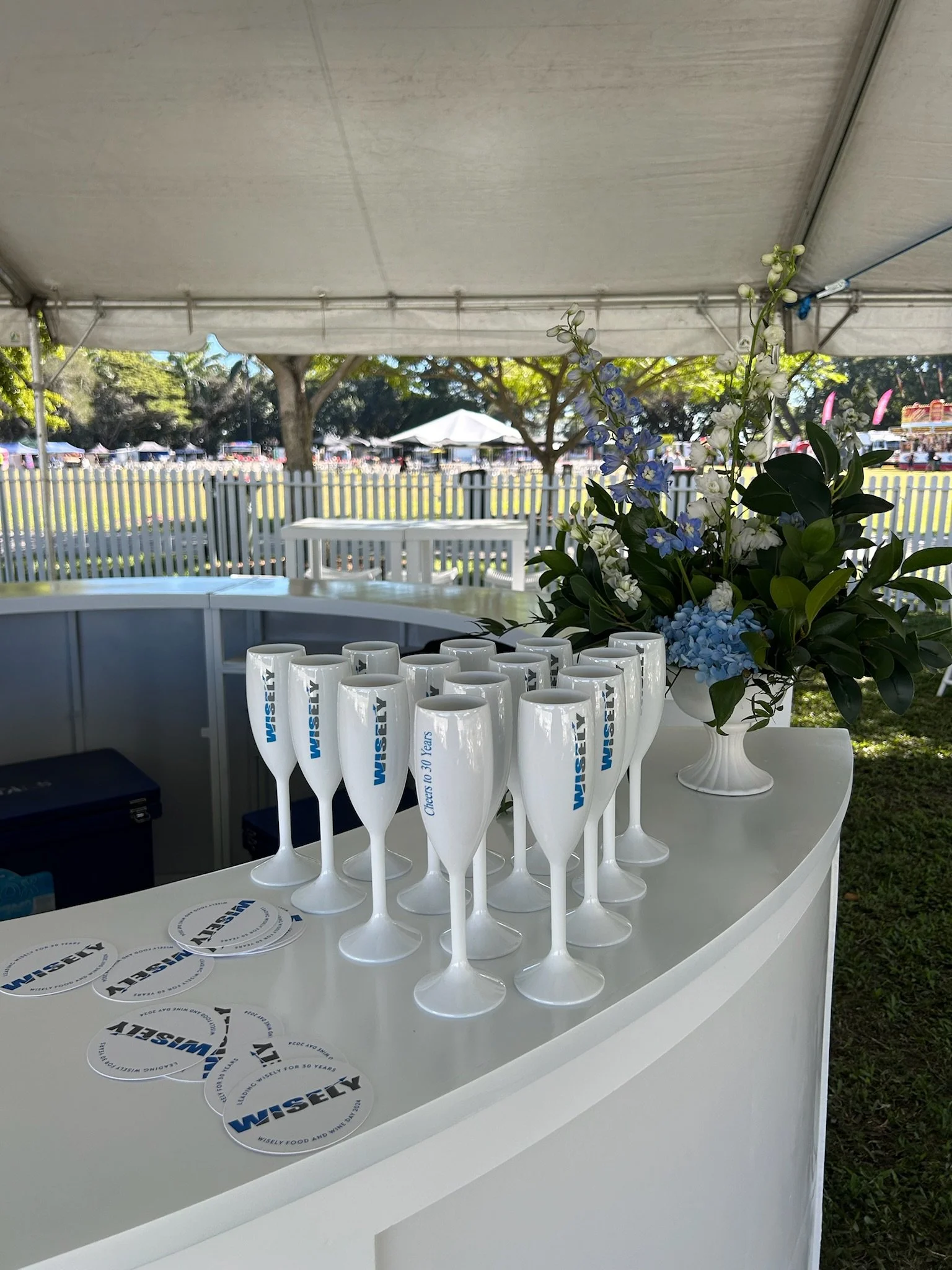 A white curved counter with Wisely branded champagne flutes and Wisely coasters, decorated with a white vase of assorted blue and white flowers, set under a tent at an outdoor event with trees, fencing, and booths in the background.
