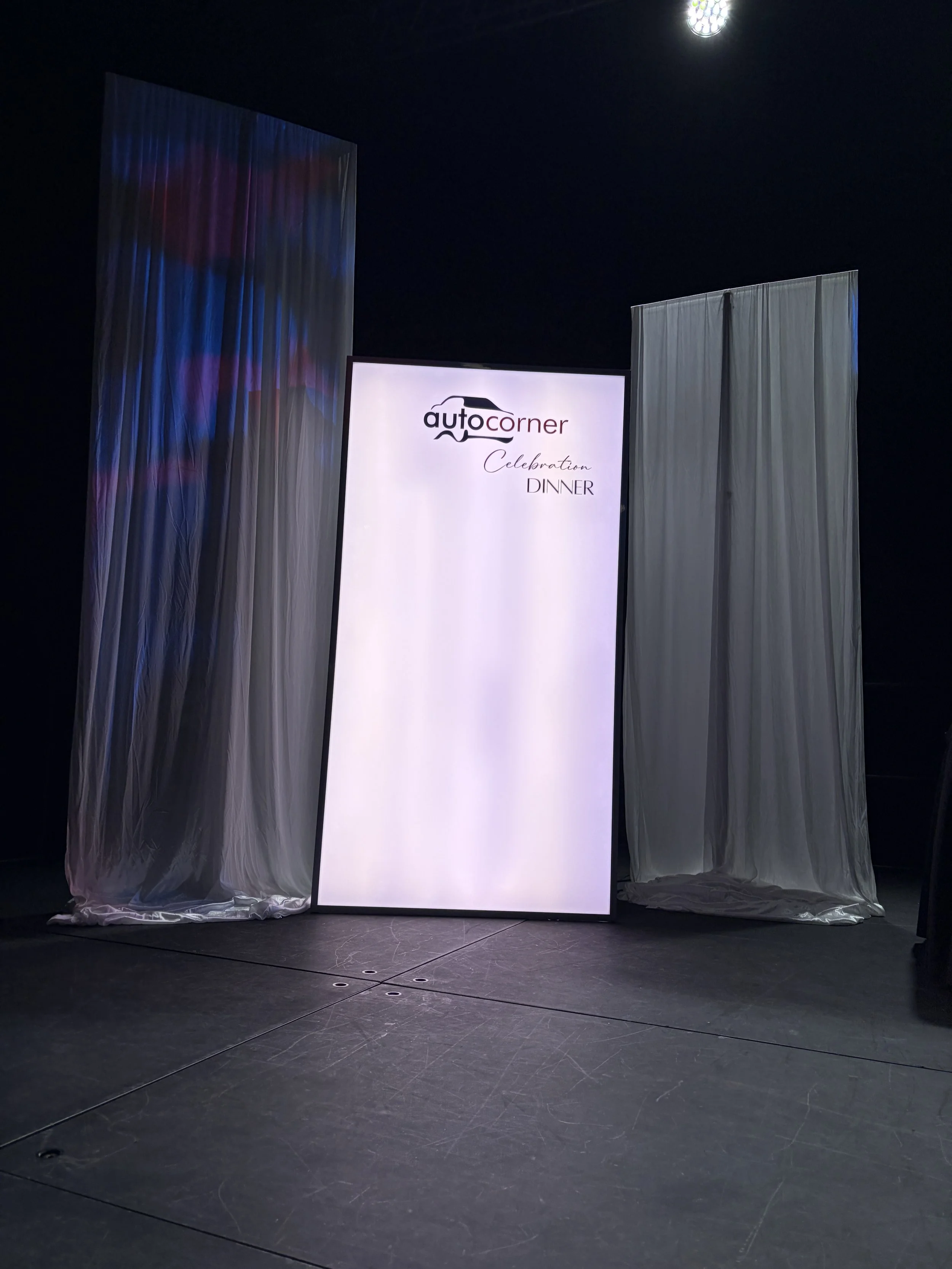A large illuminated screen displays the logo 'auto corner' with the words 'Celebration Dinner' underneath, flanked by white curtains in a dark room.