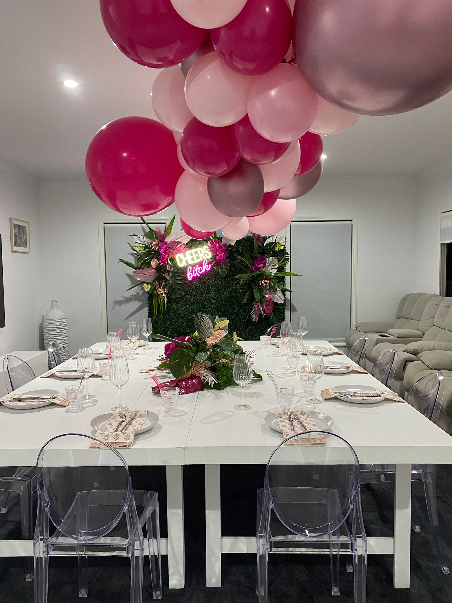 A decorated dining table with flowery centerpiece and a large cluster of pink, red, gray, and light pink balloons overhead. A neon sign on the wall says 'CHEERS bitch.' The table is set with glassware, plates, and napkins, with transparent chairs aro