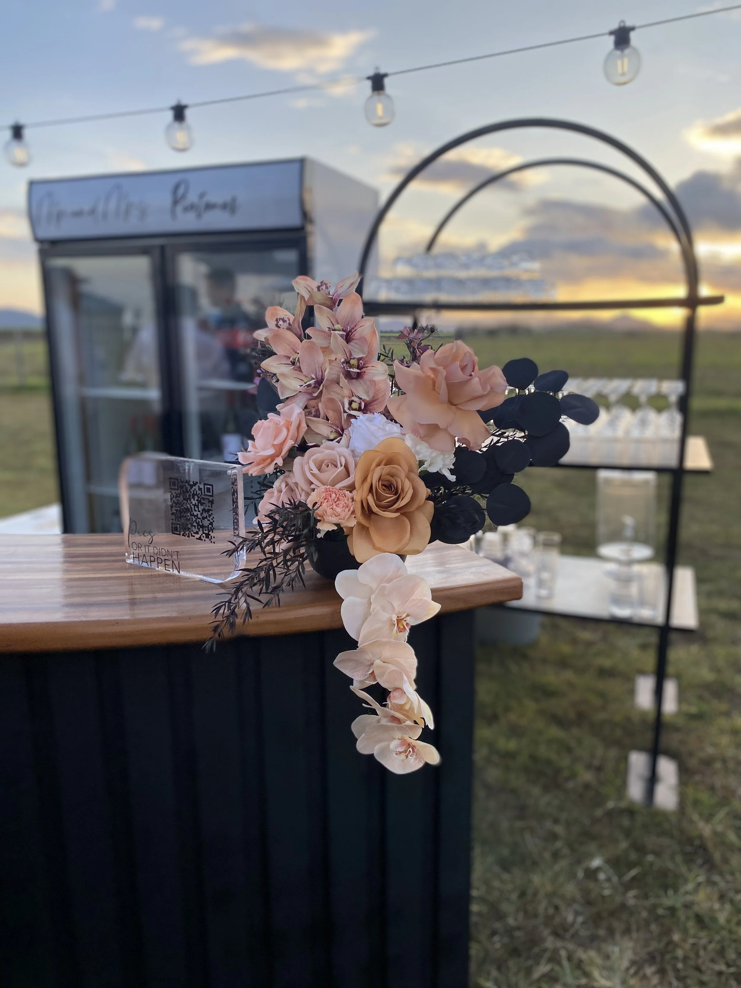 Floral arrangement with pink and peach roses and orchids on a wooden counter at an outdoor event during sunset, with a black metal structure and a refrigerator in the background.