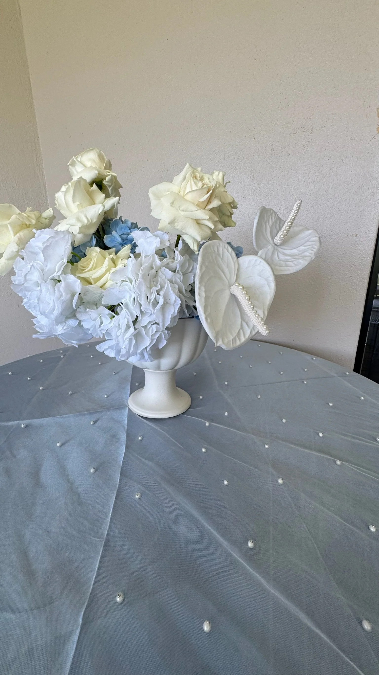White floral arrangement with roses, hydrangeas, and decorative anthuriums on a table with a sheer tablecloth decorated with small pearls.