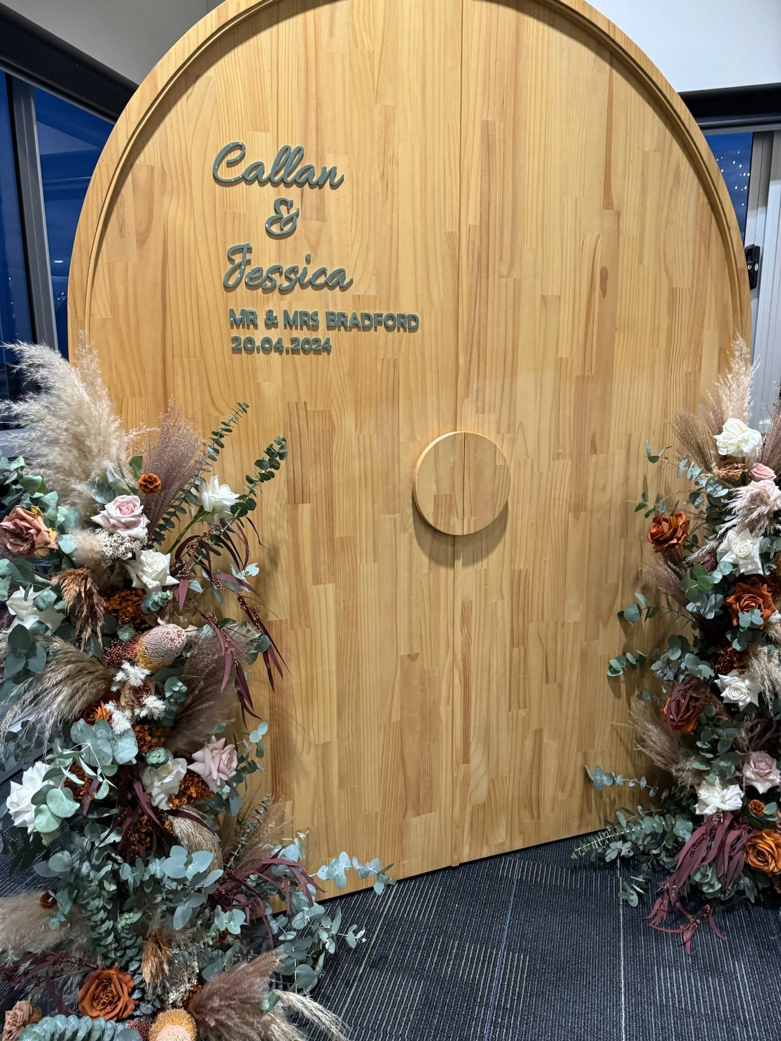 Wooden wedding backdrop with floral arrangements on both sides, inscribed with names Callan & Jessica, date 20.04.2024, and title Mr & Mrs Bradford.