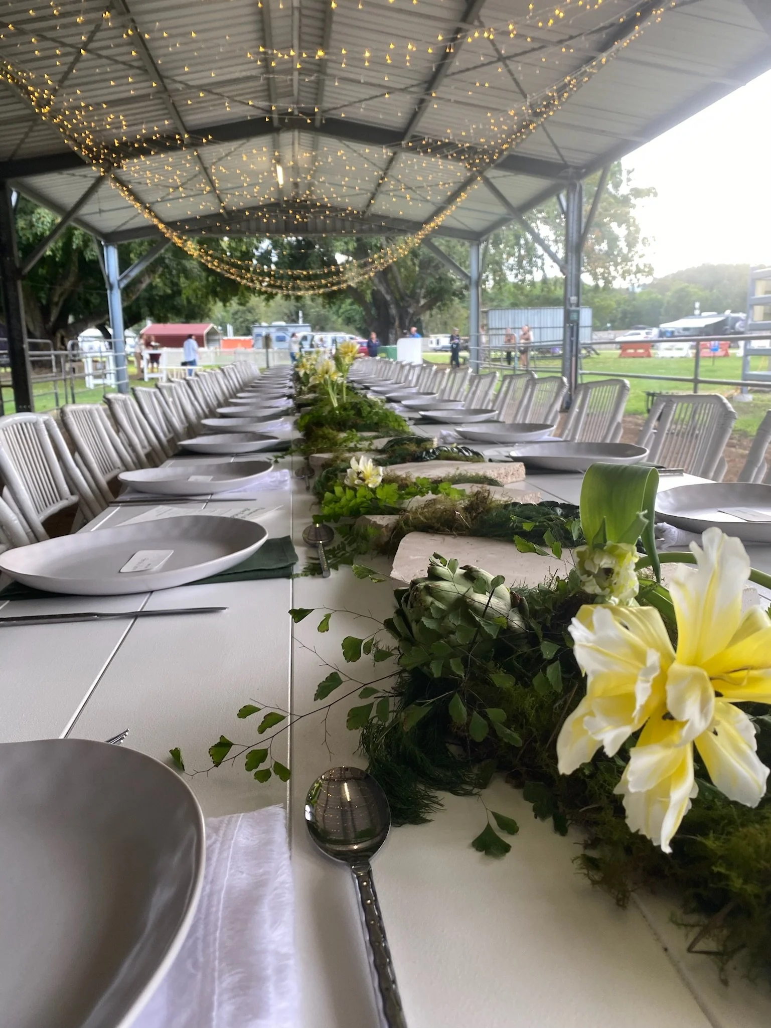 Long banquet table set for an event under a metal pavilion with string lights overhead, decorated with a central floral arrangement of white lilies and greenery.