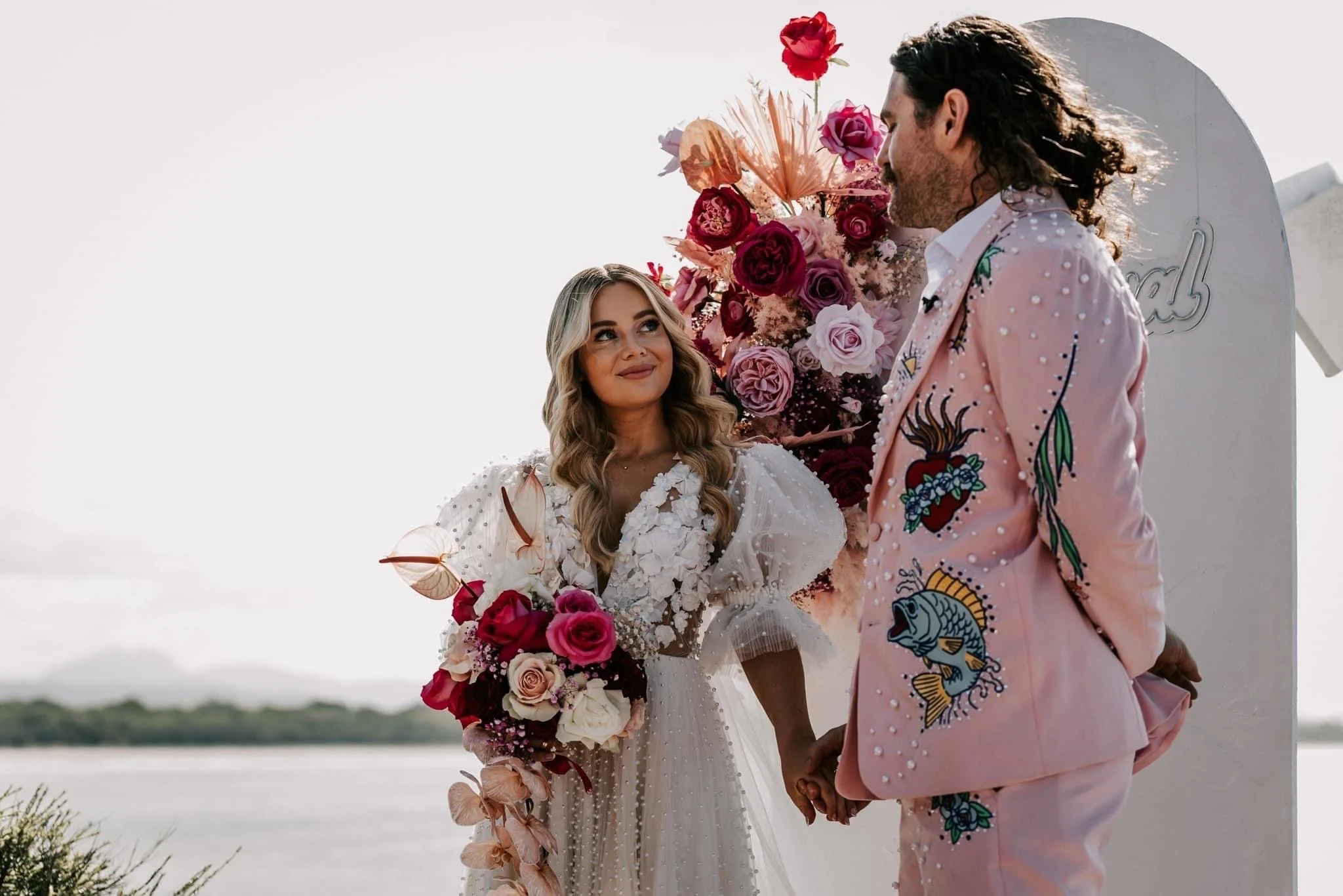 A couple getting married outdoors, holding hands, with a backdrop of colorful flowers and a white structure.