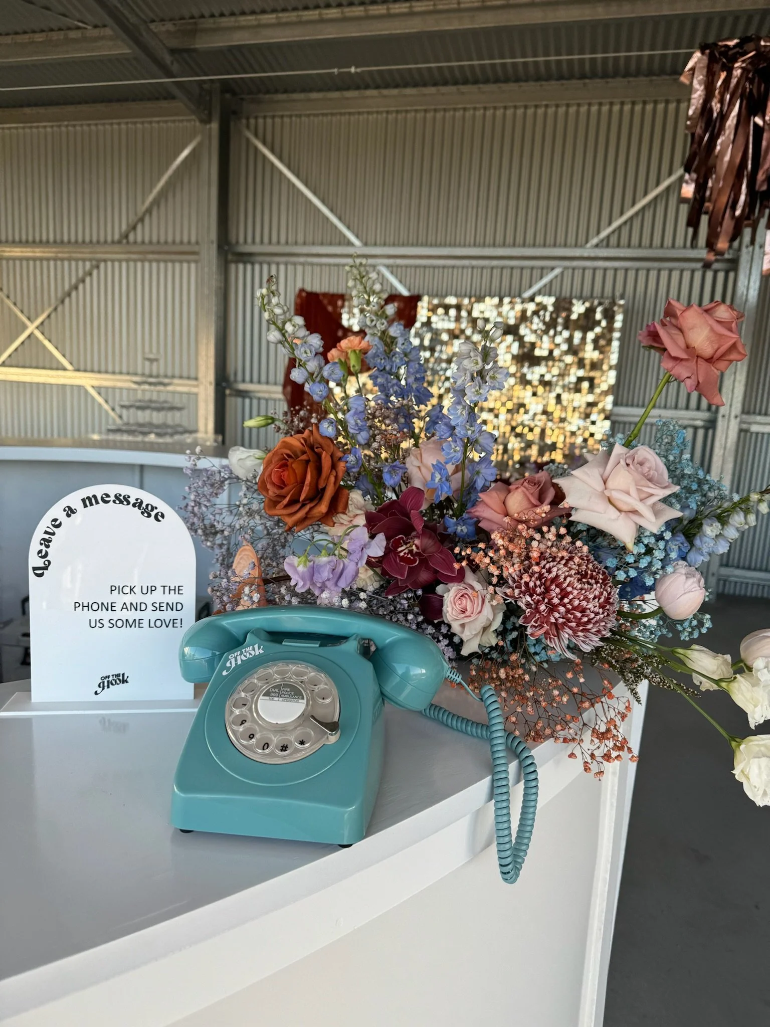 A vintage teal rotary telephone placed on a white table next to a colorful floral arrangement with roses, delphiniums, and other flowers, in front of a sign that reads 'leave a message' and 'pick up the phone and send us some love!'