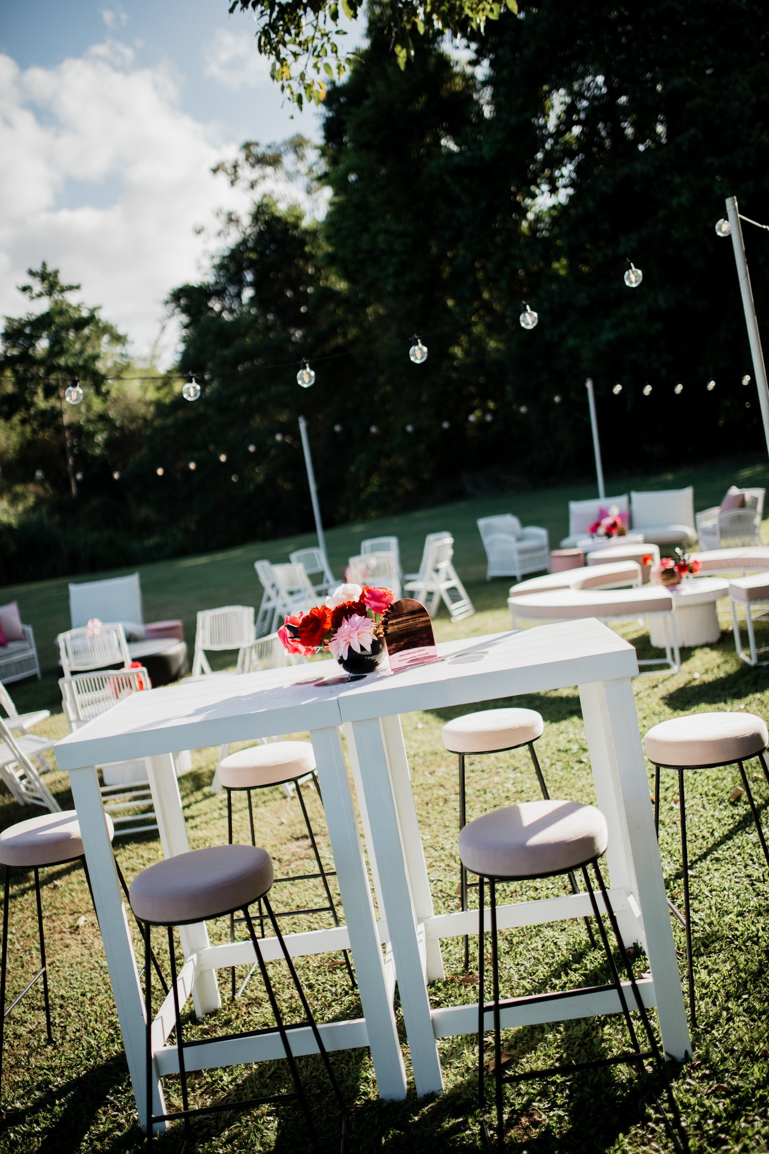 Outdoor party setup with white high-top table, pink and red flower arrangement, and various white chairs and seating areas under string lights, on a grassy yard with trees and a partly cloudy sky.