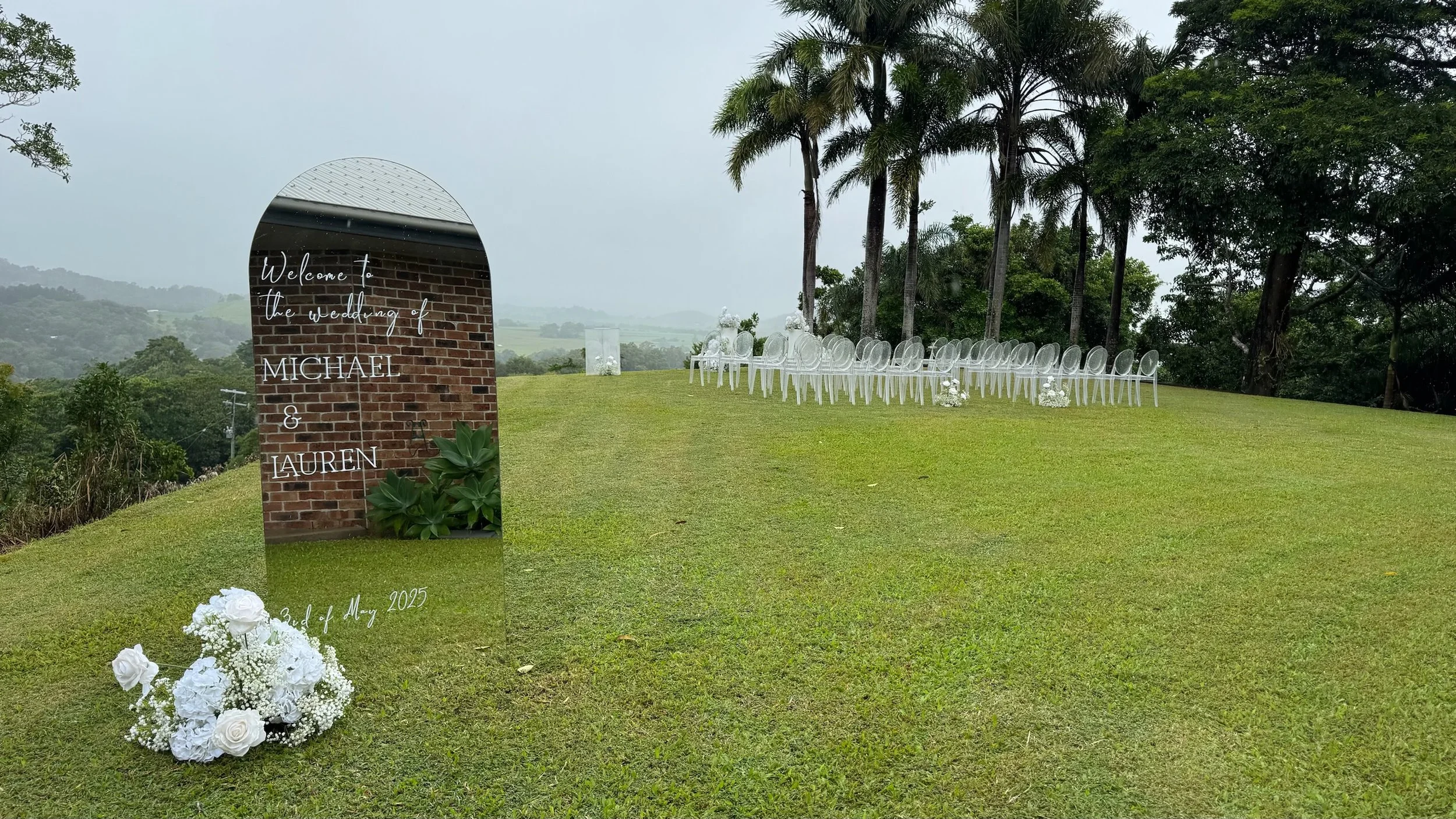 Outdoor wedding ceremony setup on a grassy hill with transparent chairs lined up, white flowers, and trees in the background; a large mirror with wedding details and floral arrangements in the foreground.