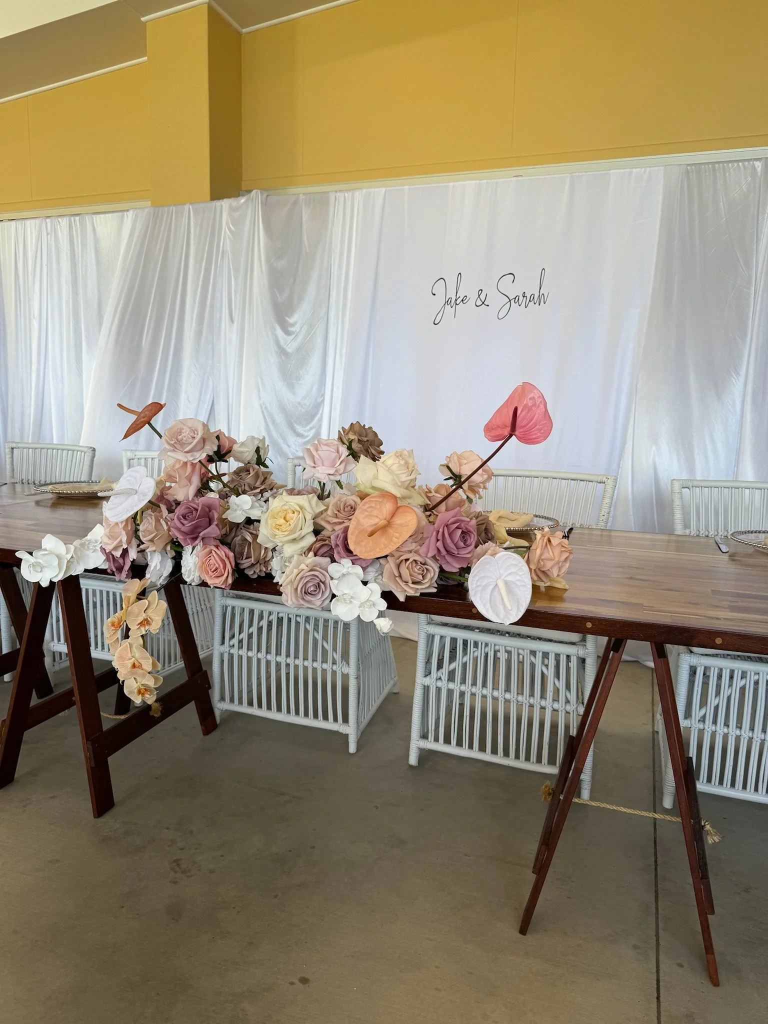 Table decorated with pastel-colored roses and anthuriums, set up for a wedding or celebration, with white wicker chairs and a backdrop with the names Jake & Sarah.