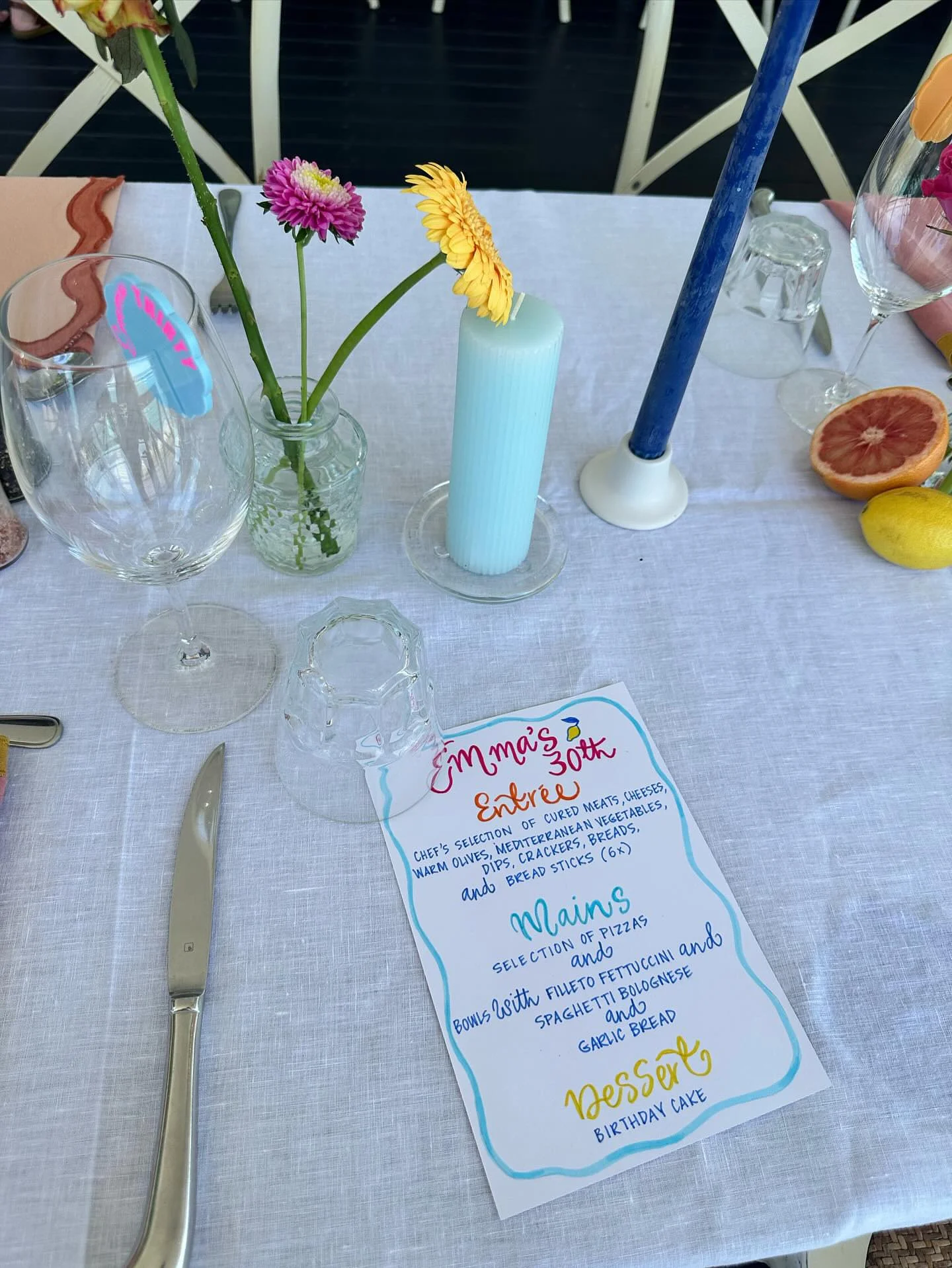 A dining table with a white tablecloth set for a celebration. In the center, there's a small vase with pink and yellow flowers, and a tall blue candle. There are wine glasses, a menu card, cutlery, and fresh citrus fruits, including grapefruit and le
