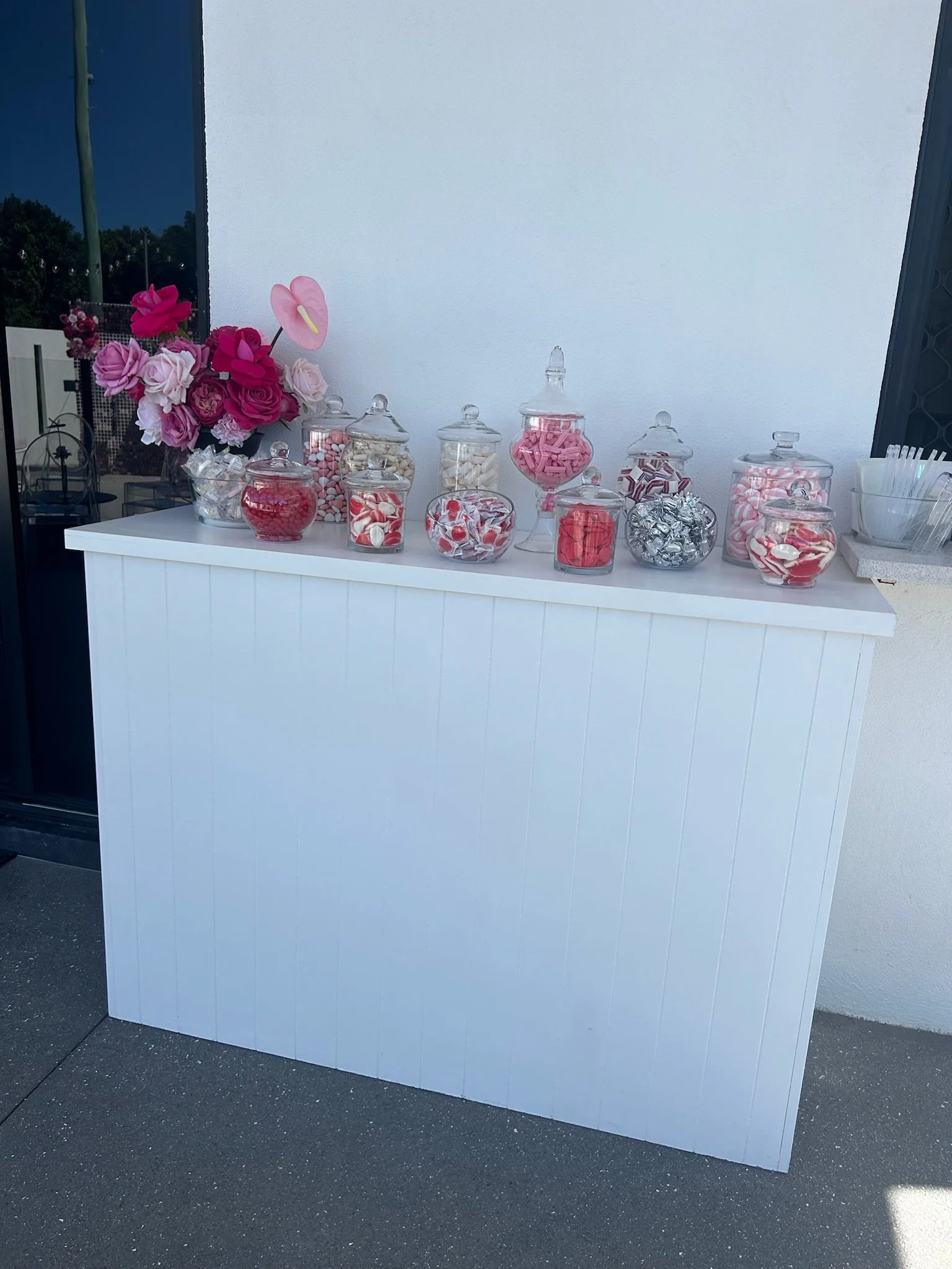 Candy display table with pink, red, and white candies in glass jars, decorated with pink and red flowers on the side.