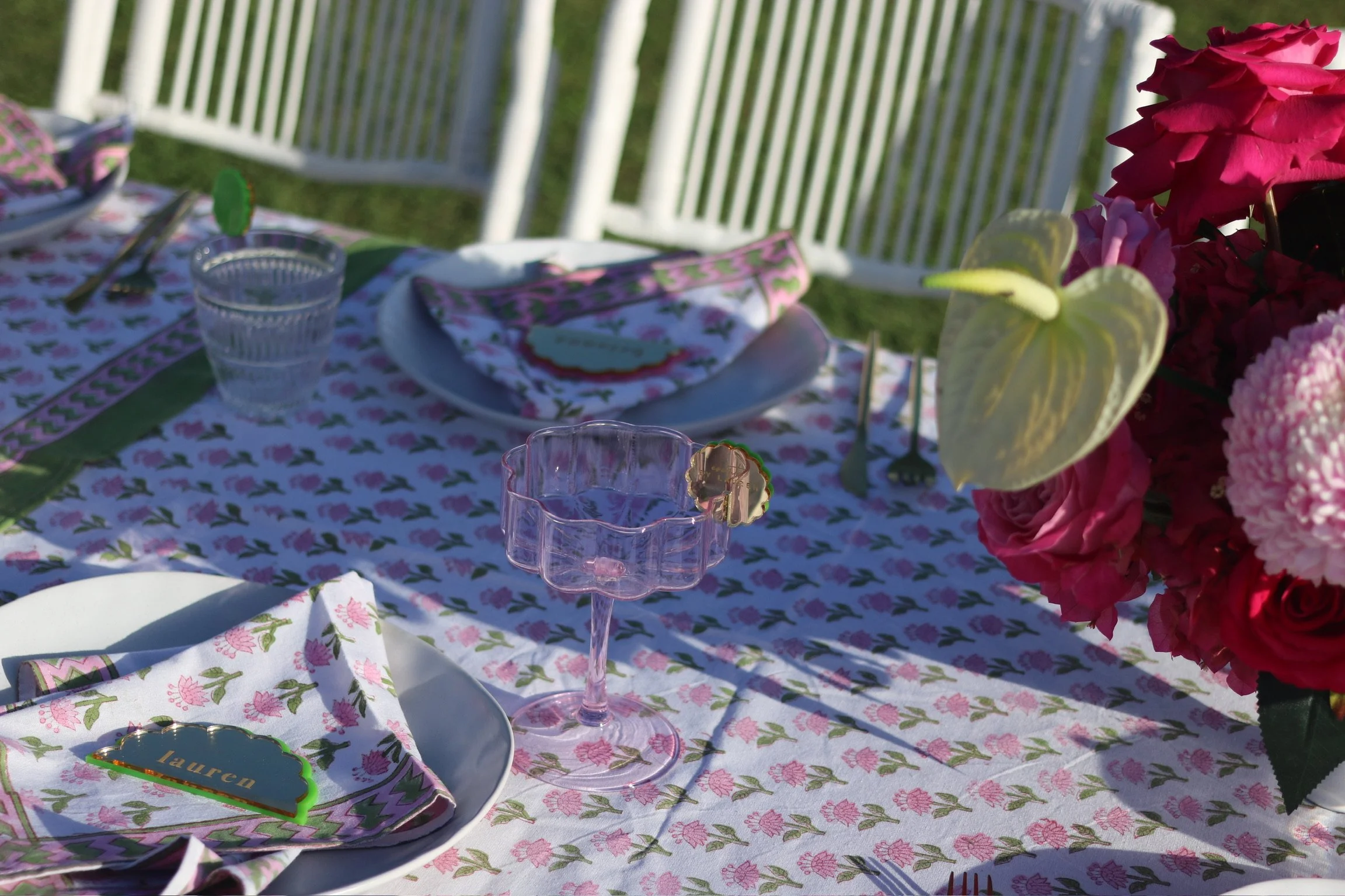 A beautifully set outdoor table with floral-patterned tablecloth, pink napkins, clear glassware, and a centerpiece of pink and white flowers, illuminated by natural sunlight.