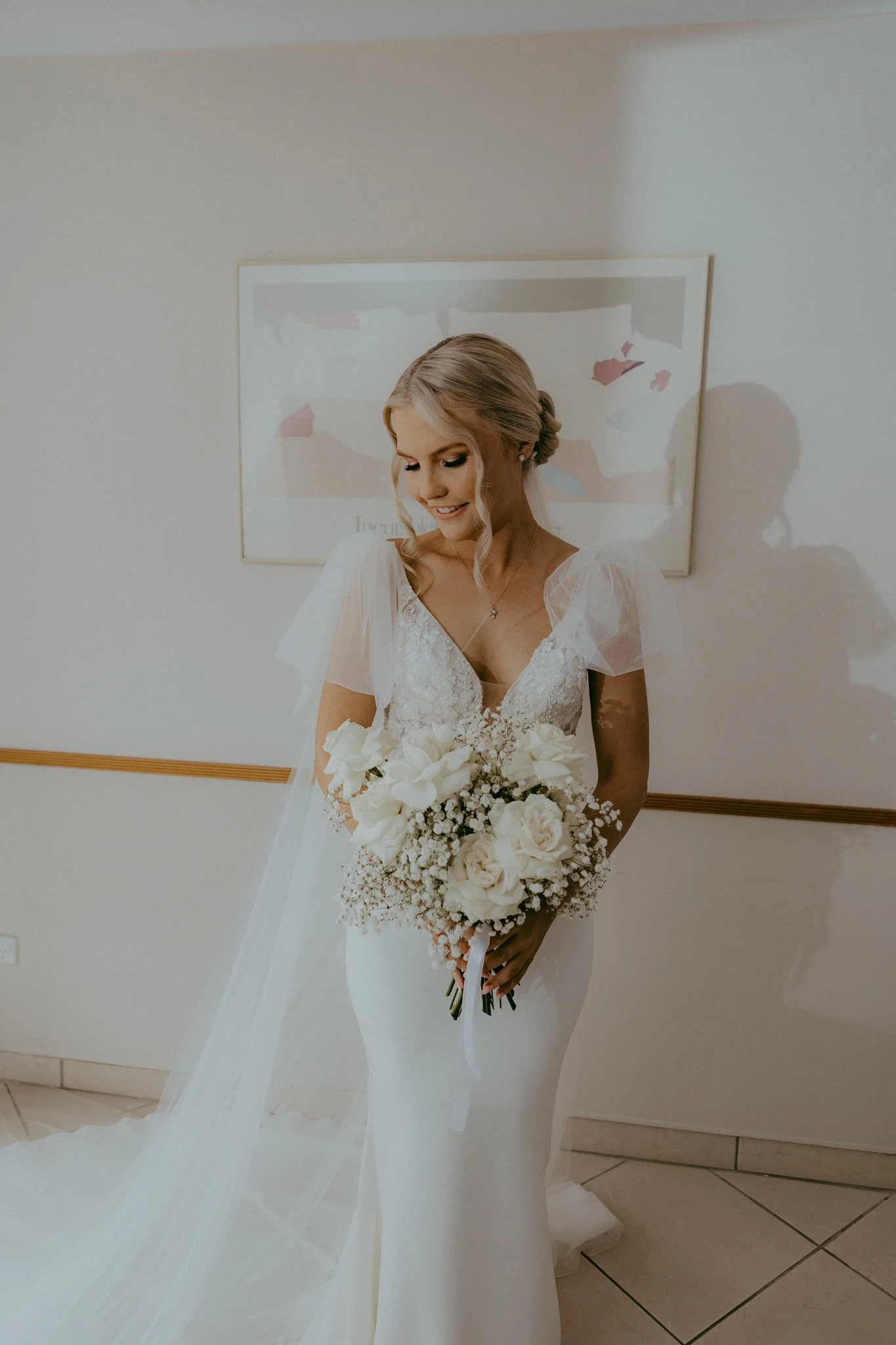 Bride in a white wedding dress holding a bouquet of white roses and baby's breath, standing indoors near a wall with a framed abstract artwork.