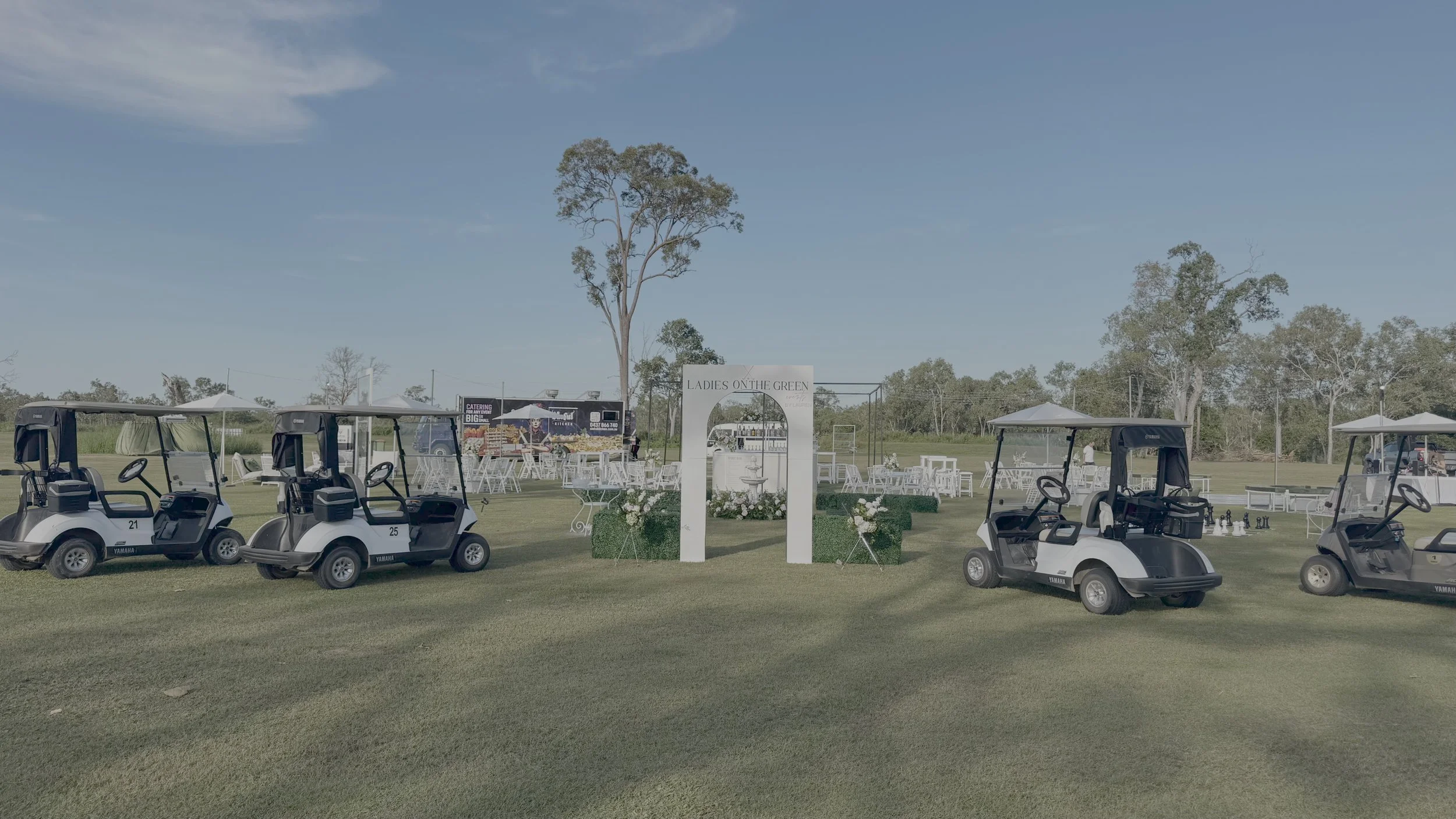 Golf carts parked on a grassy field with a white decorative archway and tables with umbrellas in the background, suggesting an outdoor event or gathering.