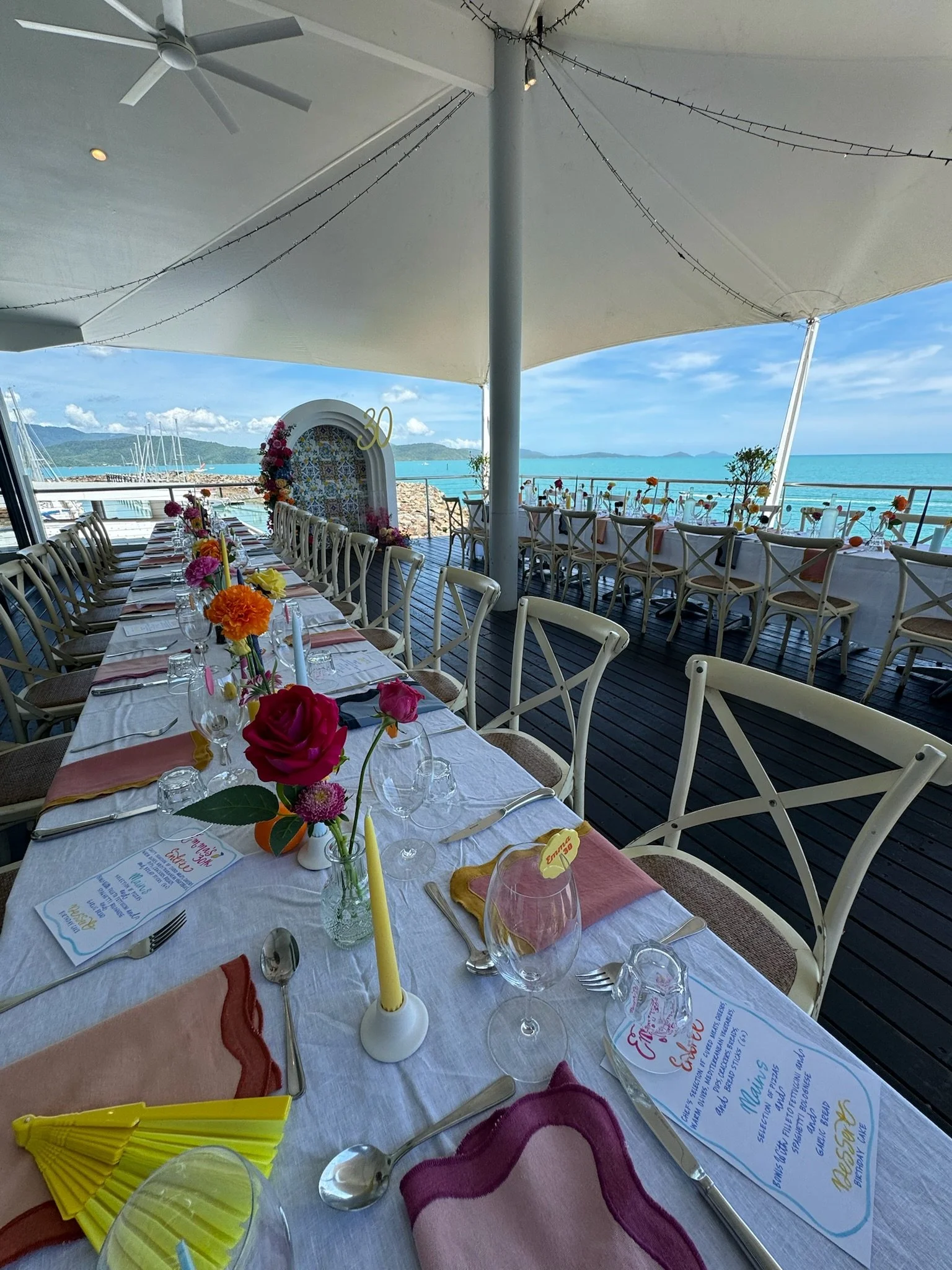 A seaside restaurant decorated for a celebration with long tables set with colorful flowers, candles, and menus, overlooking the ocean and mountains in the distance under a large white canopy.